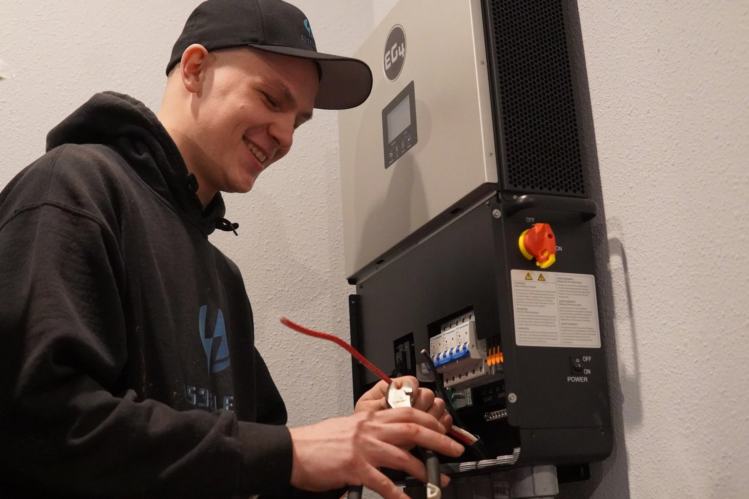 Young man working on an electrical panel mounted on a wall, smiling and wearing a black hoodie and cap.