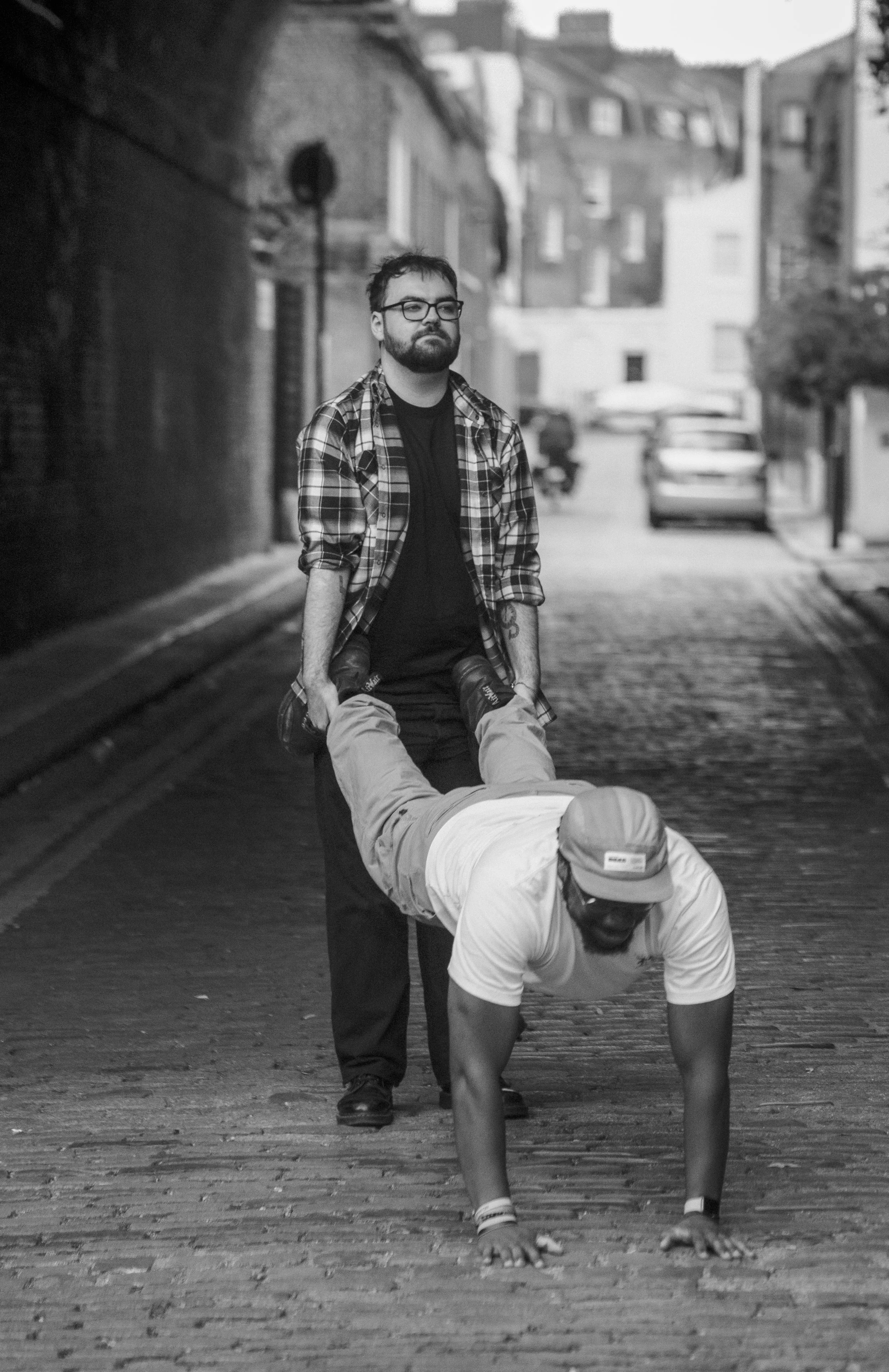 A man with glasses and a beard is holding onto a person's ankles while they are walking on hands on a cobblestone street in an urban area, with buildings and parked cars in the background.