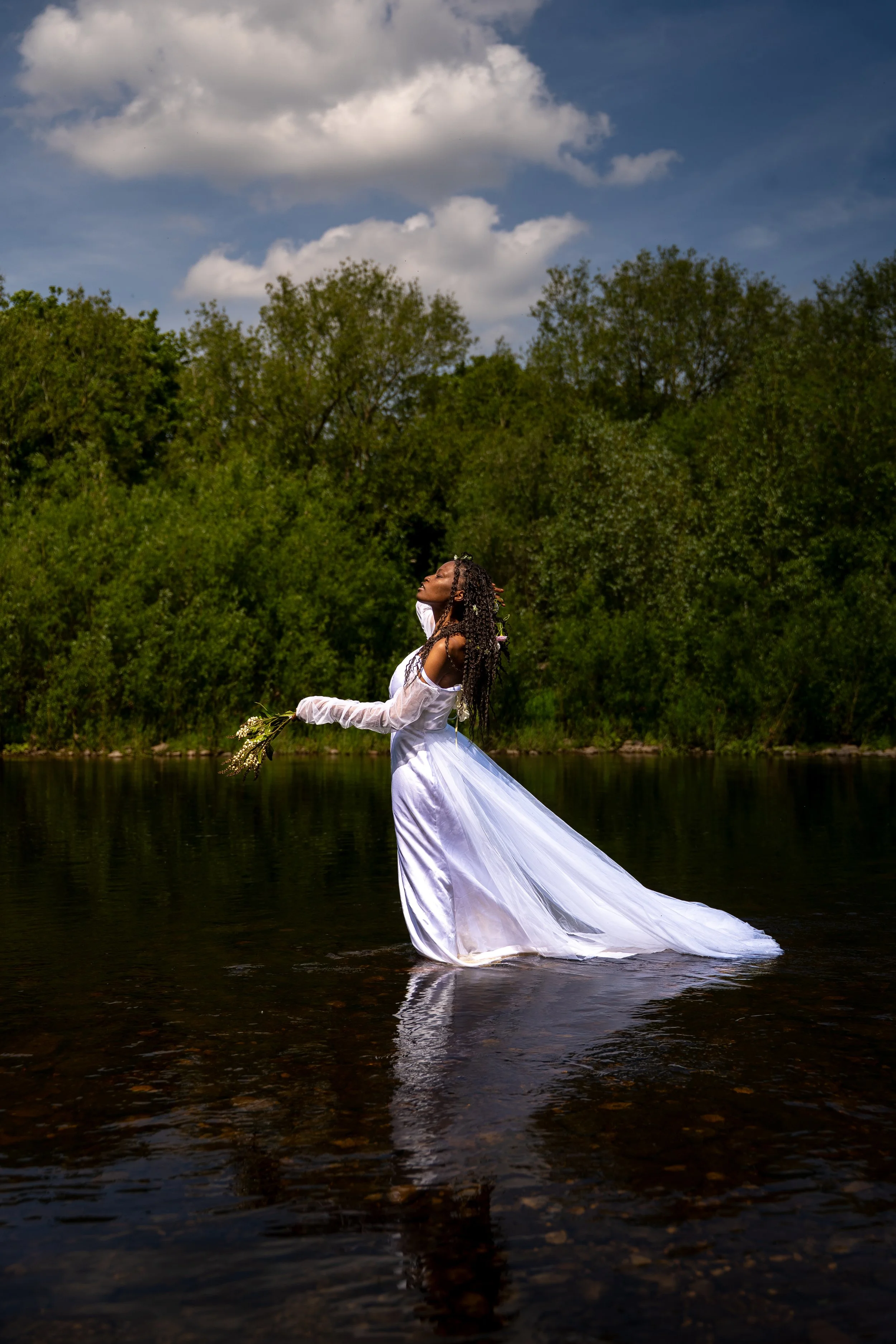 A woman in a flowing white dress stands in shallow water, holding a small bouquet of flowers, with her head tilted back and eyes closed, surrounded by greenery and under a partly cloudy sky.