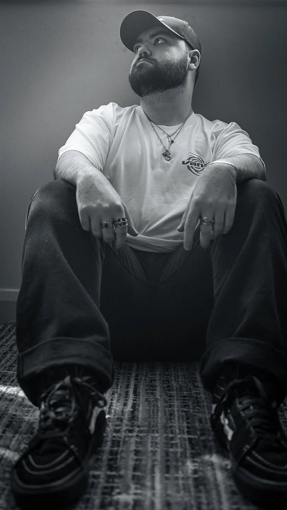 A man with a beard and cap sitting on the floor with his legs apart, looking up and to the left. He is wearing a white T-shirt, rings, necklaces, and sneakers in a black and white photo.