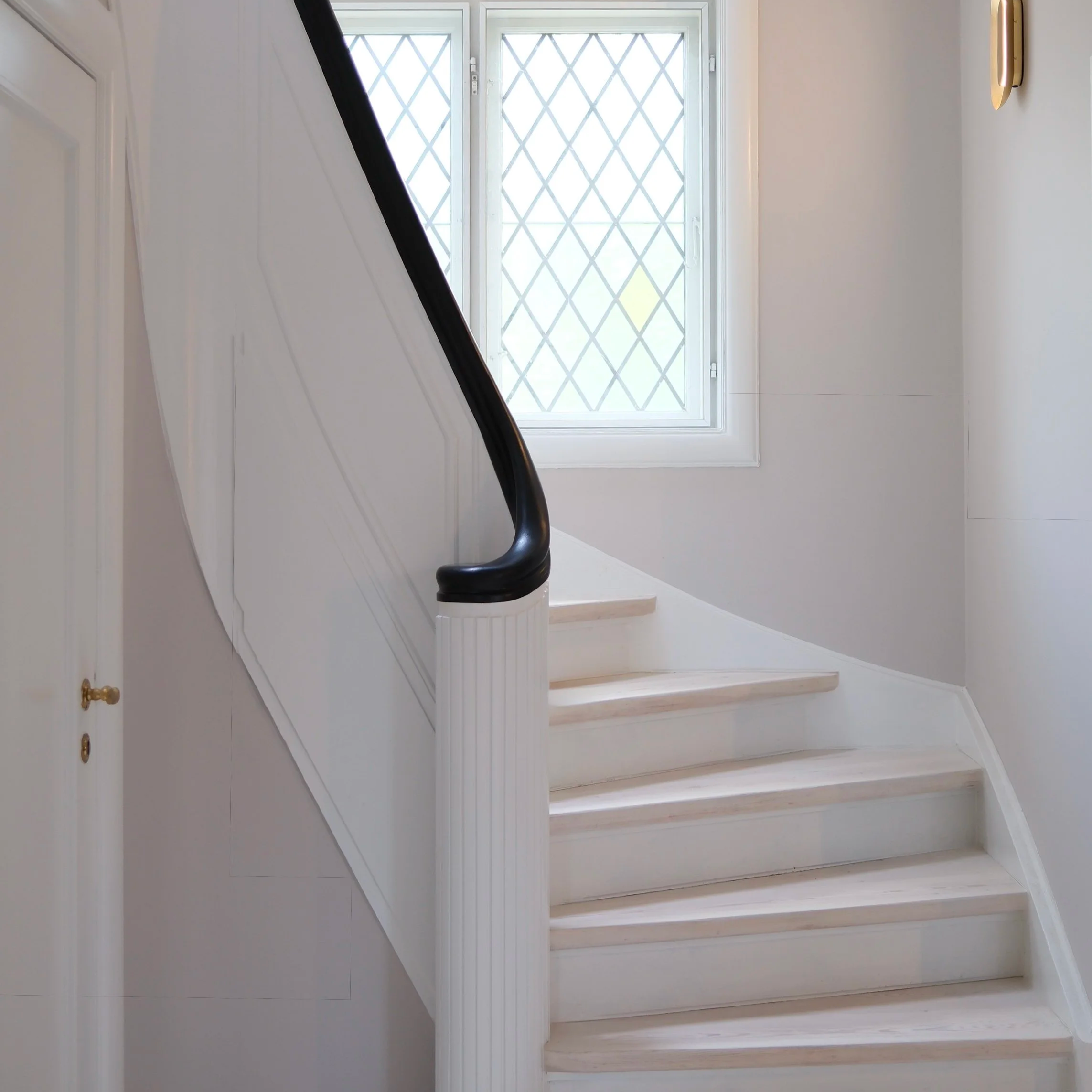 Interior view of a staircase with white stairs and black handrail, window with diamond-patterned security grille, white walls, and a wall-mounted light fixture.