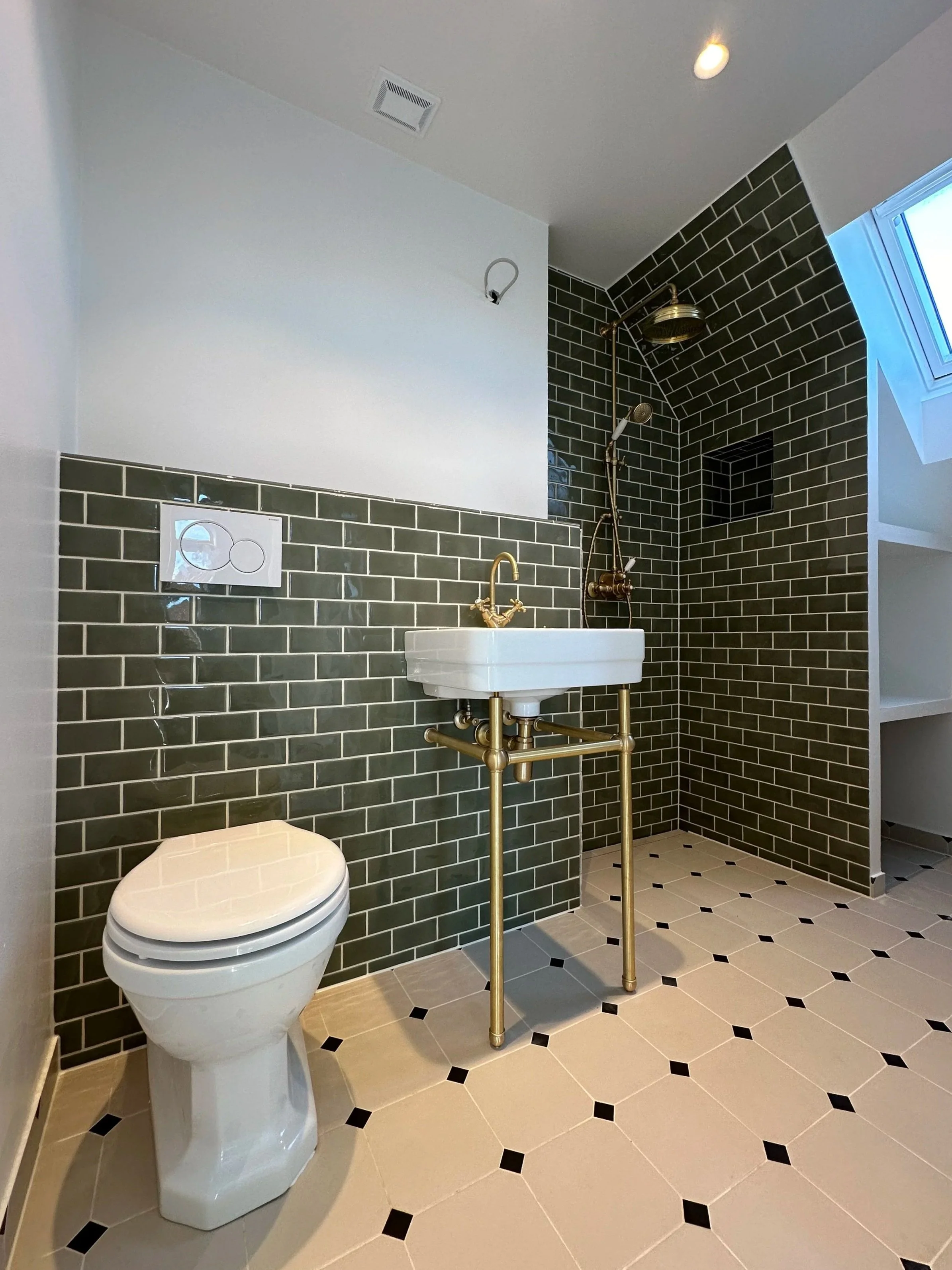 Modern bathroom with black subway tile walls, white floor tiles with black accents, a wall-mounted toilet, a small rectangular sink with gold fixtures, and a shower area with a brass rain shower head and black niche for toiletries. There is a window in the shower area and a ceiling light.