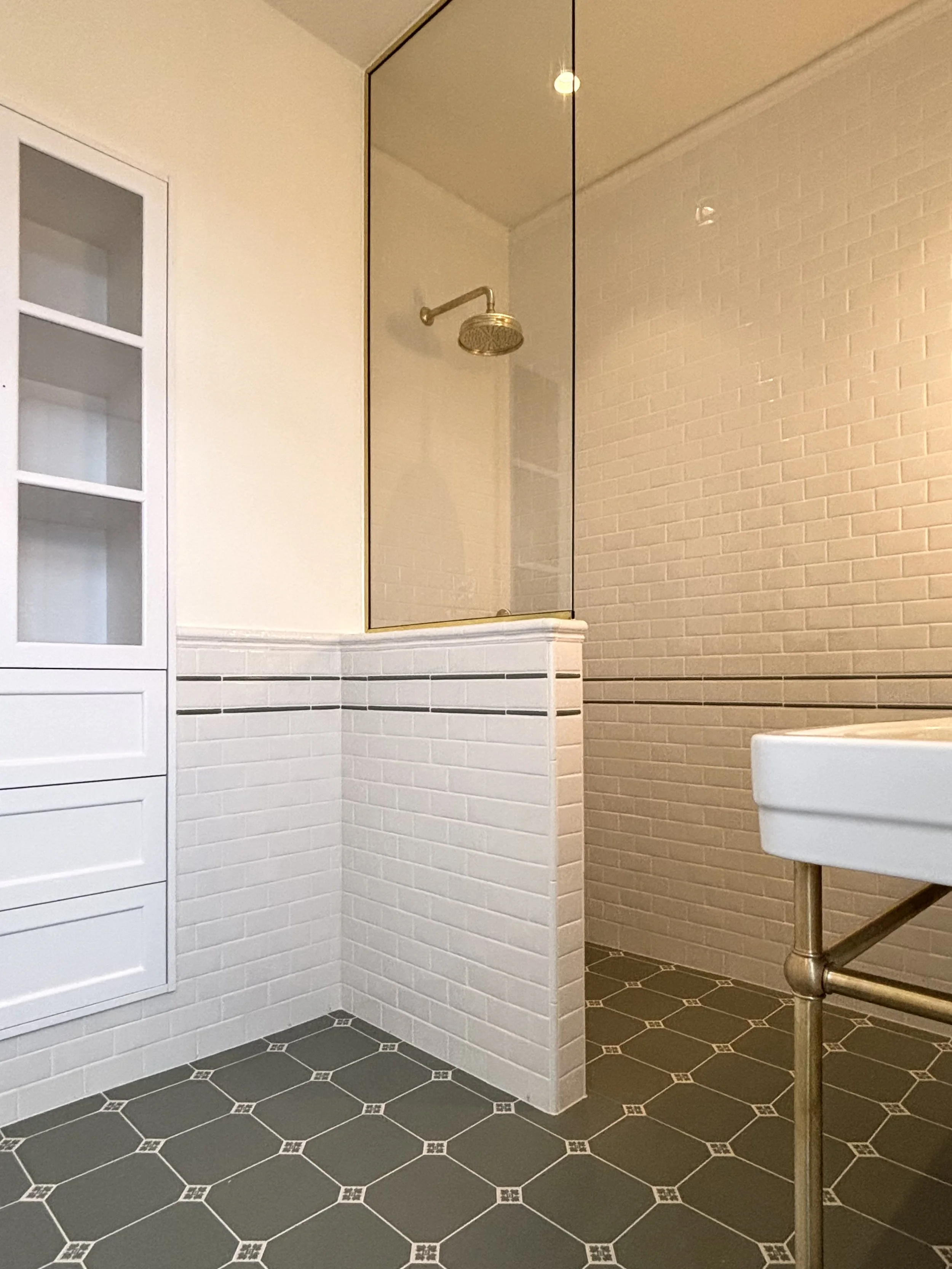 A bathroom with black and white tiled flooring, a white tiled lower wall, a white cupboard with glass panels, a glass shower enclosure with a brass showerhead, and part of a white sink with a brass pipe.