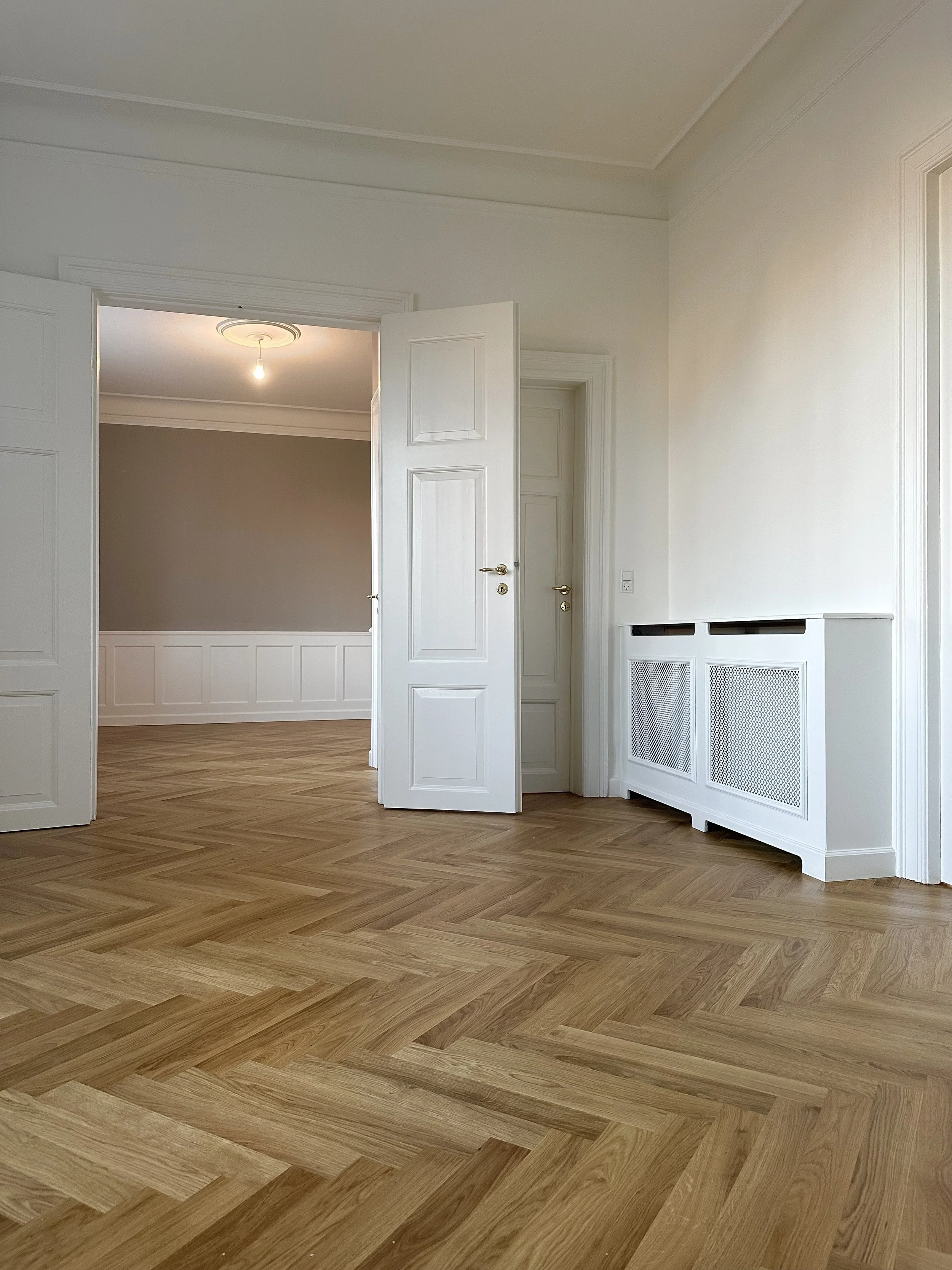 Empty room with white walls, open white doors, and a herringbone-patterned wooden floor.