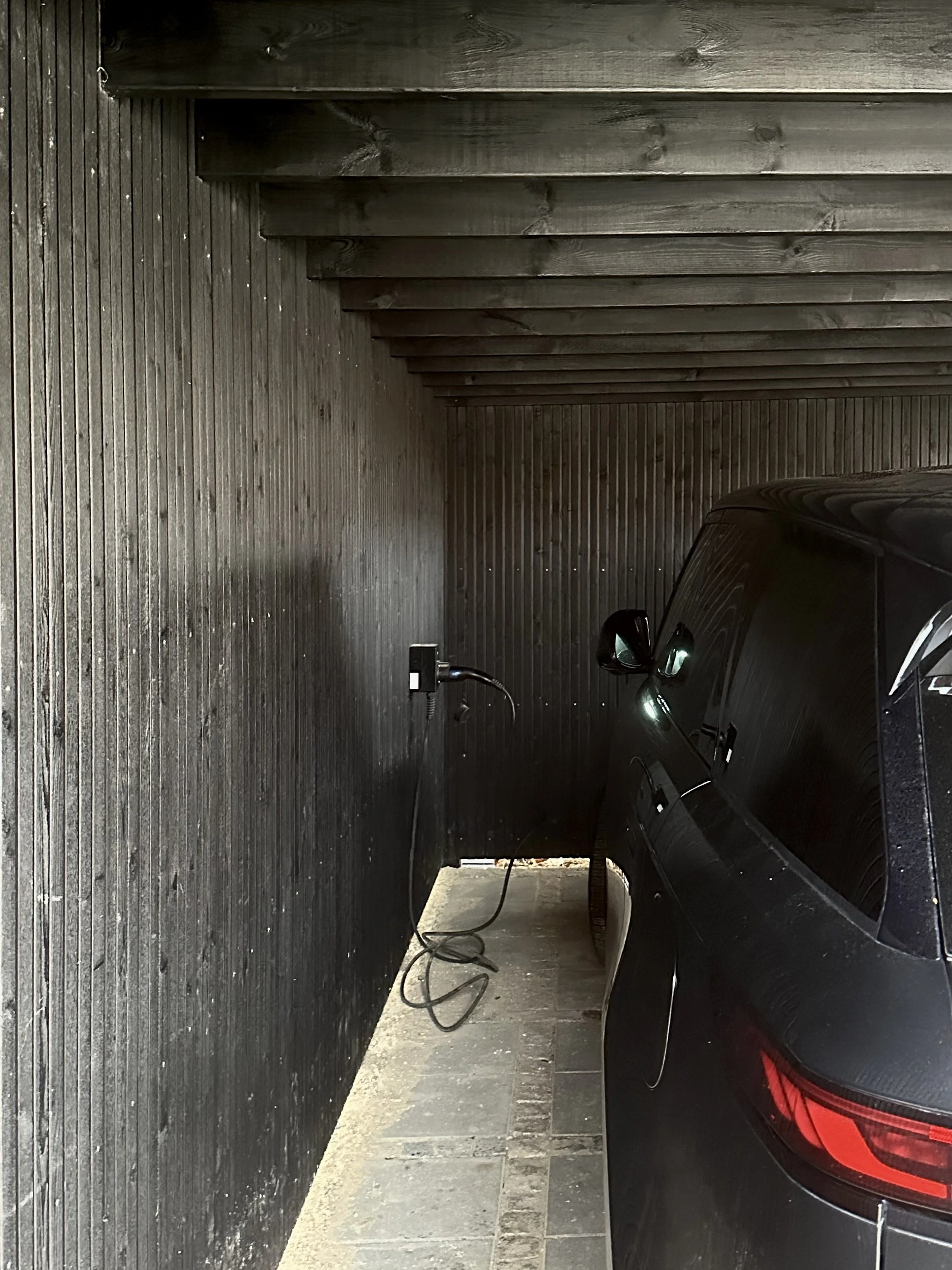 A black car parked in a narrow, covered parking space with wooden walls and ceiling, an electrical outlet on the wall, and an extension cord hanging from the outlet.