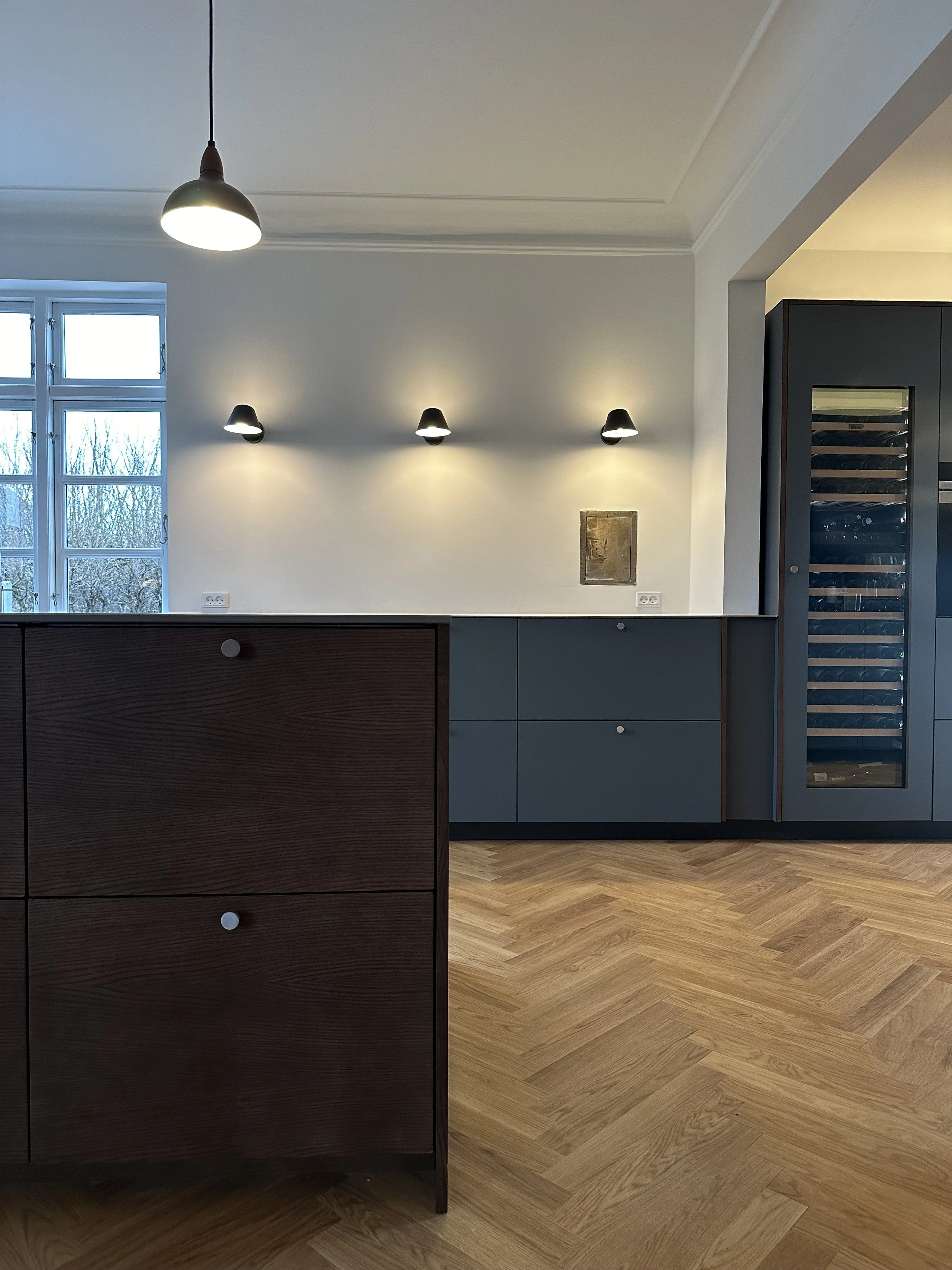 Interior of a modern room with wooden flooring, white walls, decorative molding, a window, a hanging lamp, wall sconces, and a built-in wine cooler.