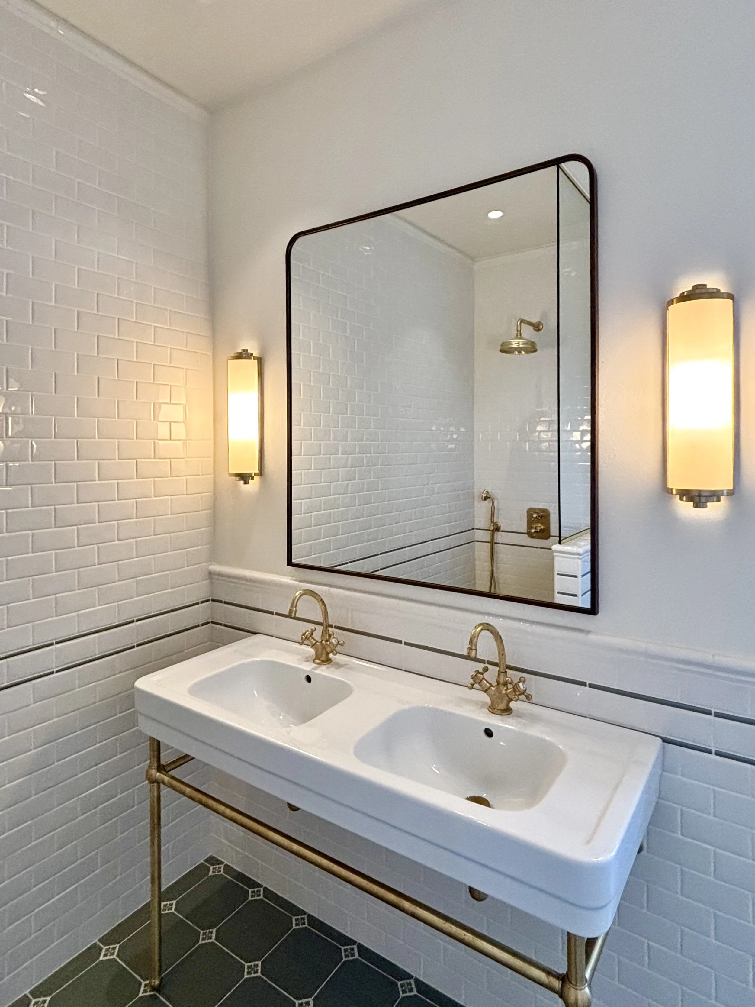 A bathroom with white subway tile walls, a large rectangular mirror, and vintage-style brass fixtures, including a double sink and wall-mounted lights.
