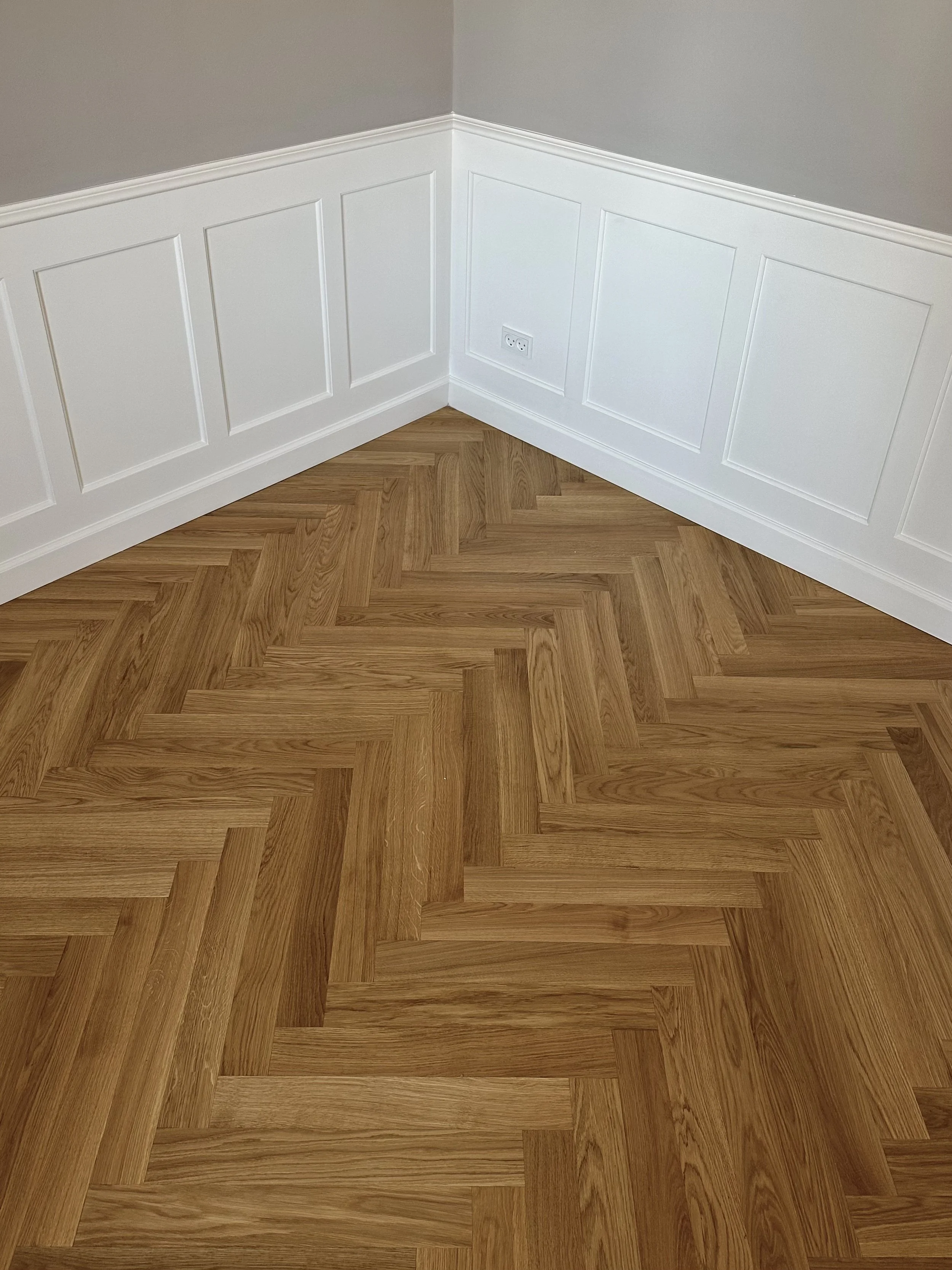 Corner of a room with gray walls, white wainscoting, and a hardwood herringbone patterned floor.