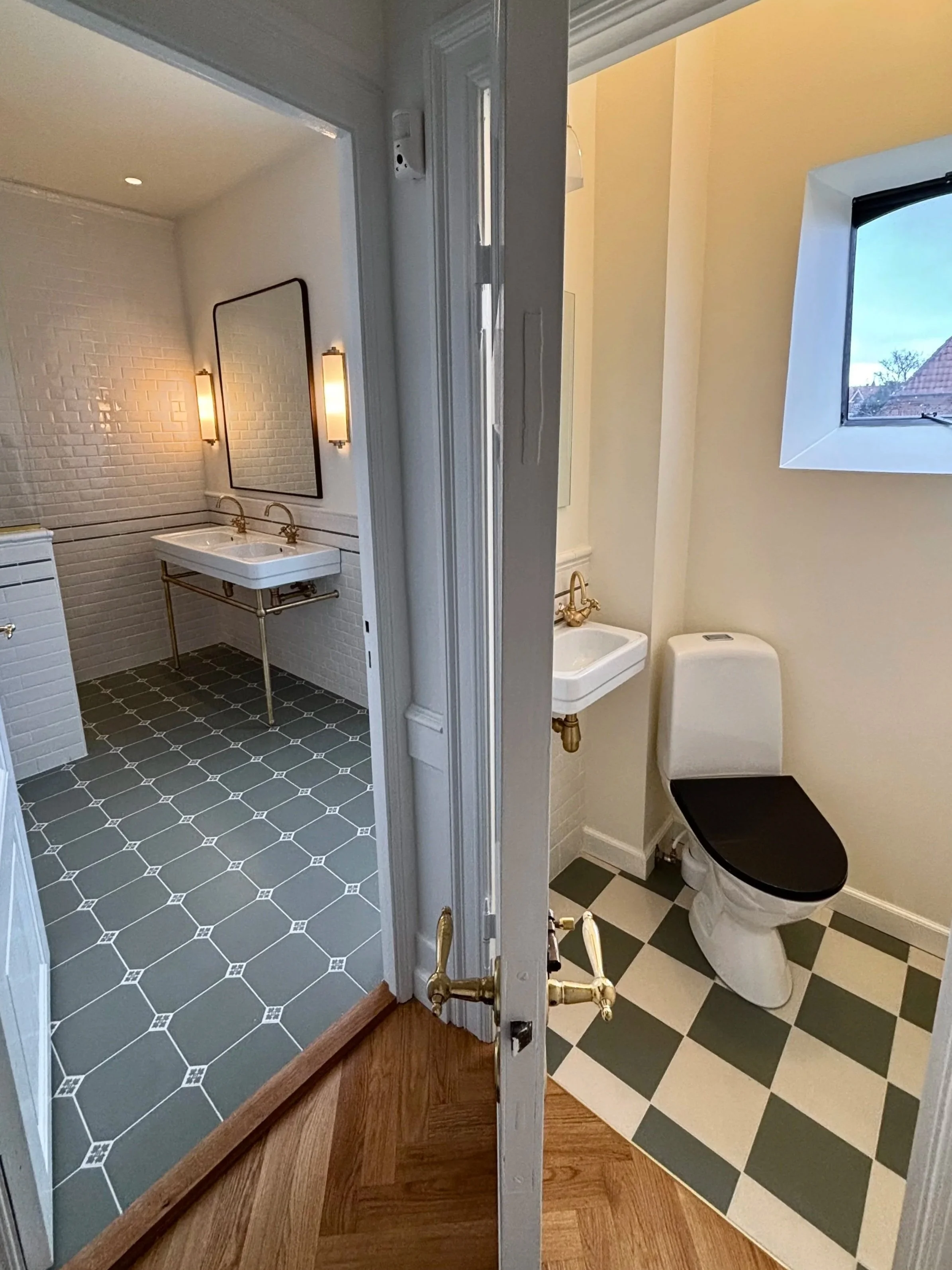 View of a bathroom with a white brick wall, double sink, gold faucet, and black patterned floor, seen through an open door with a mirror reflecting hardwood flooring from another room.