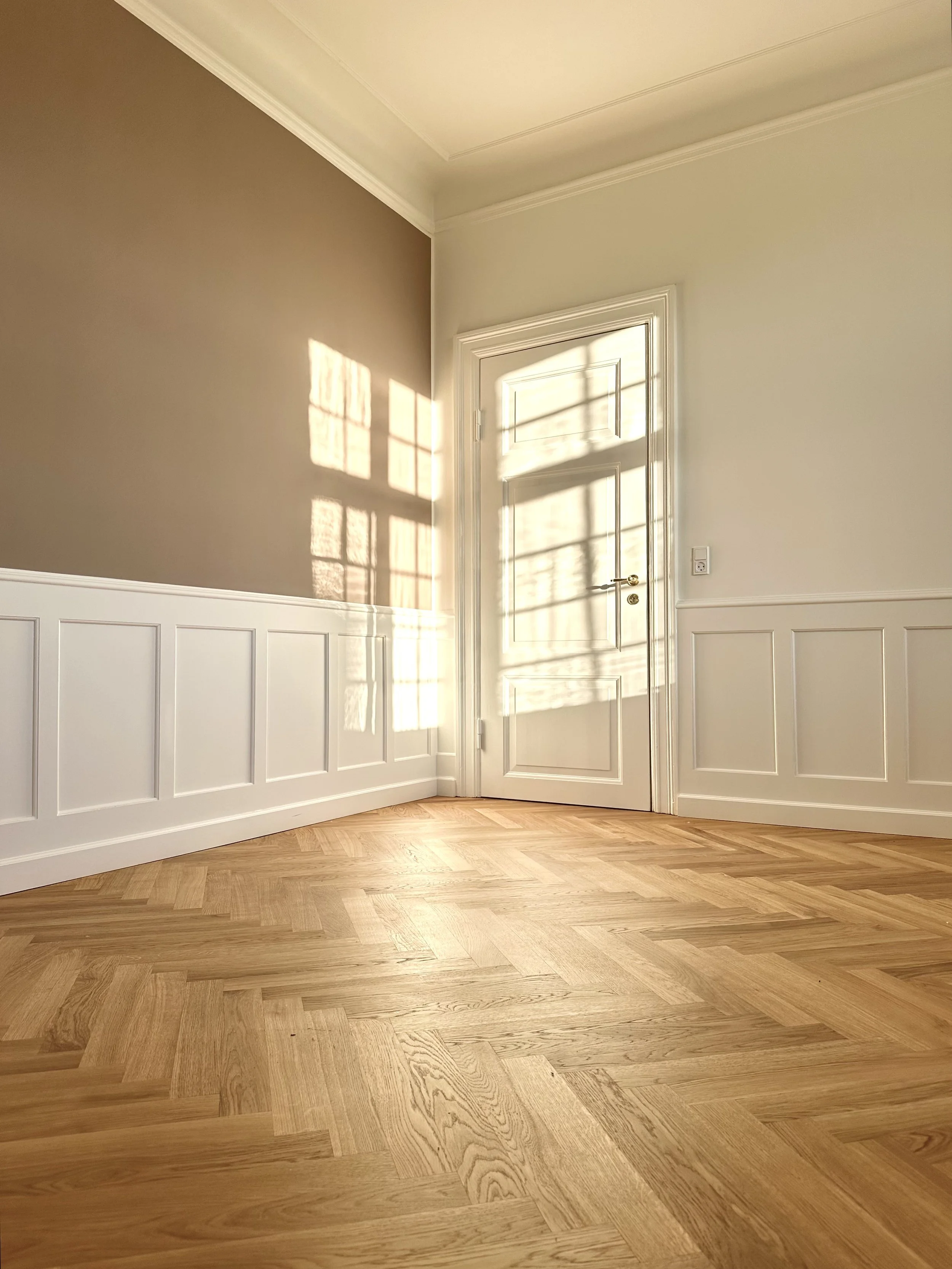 A room with a wooden herringbone floor, a beige wall on the left, white wainscoting on the lower part of the walls, a white door with shadows of window panes cast on it, and sunlight coming through the window illuminating the room.