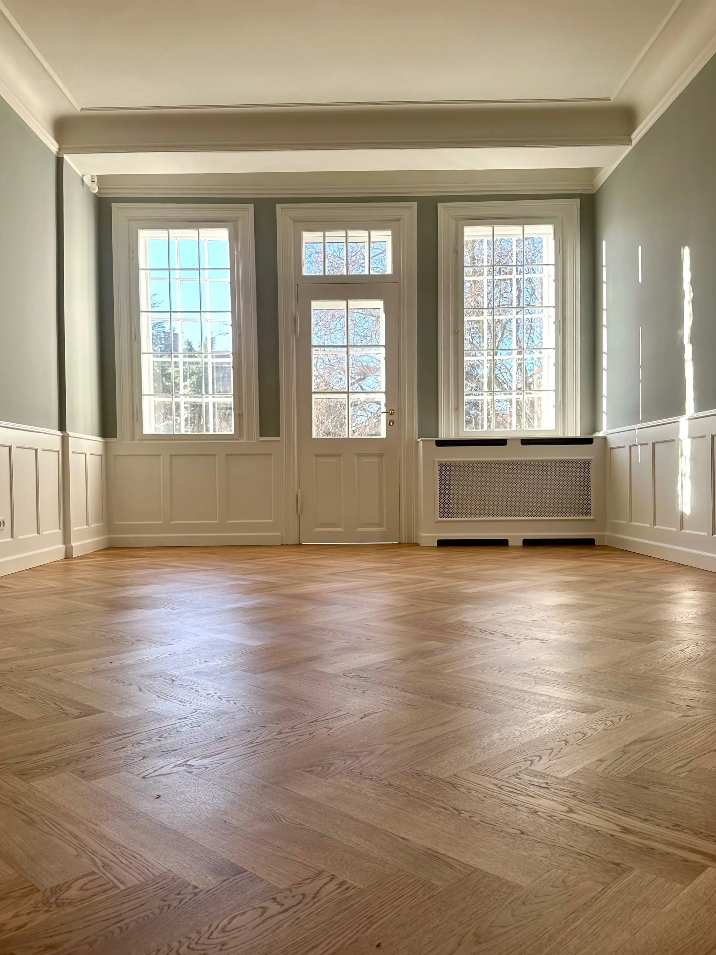 Empty room with hardwood flooring, large windows, and decorative wall paneling with a door in the center.