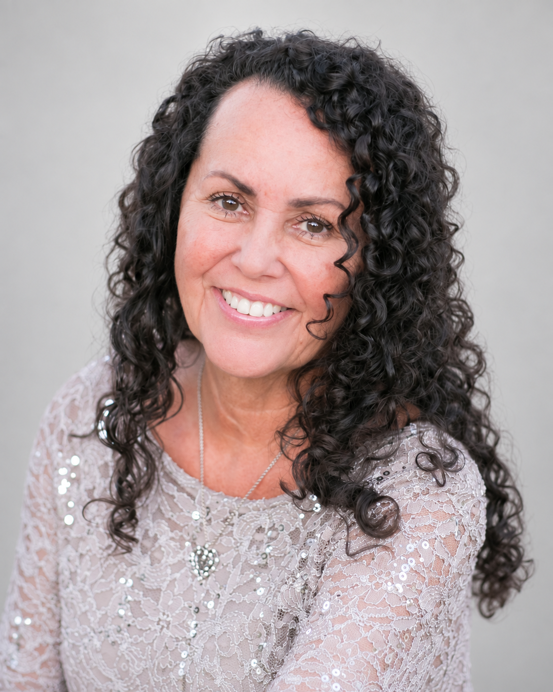 A woman with long, curly black hair, smiling outdoors in a beige lace dress with sequins, standing in front of a blurred green background.