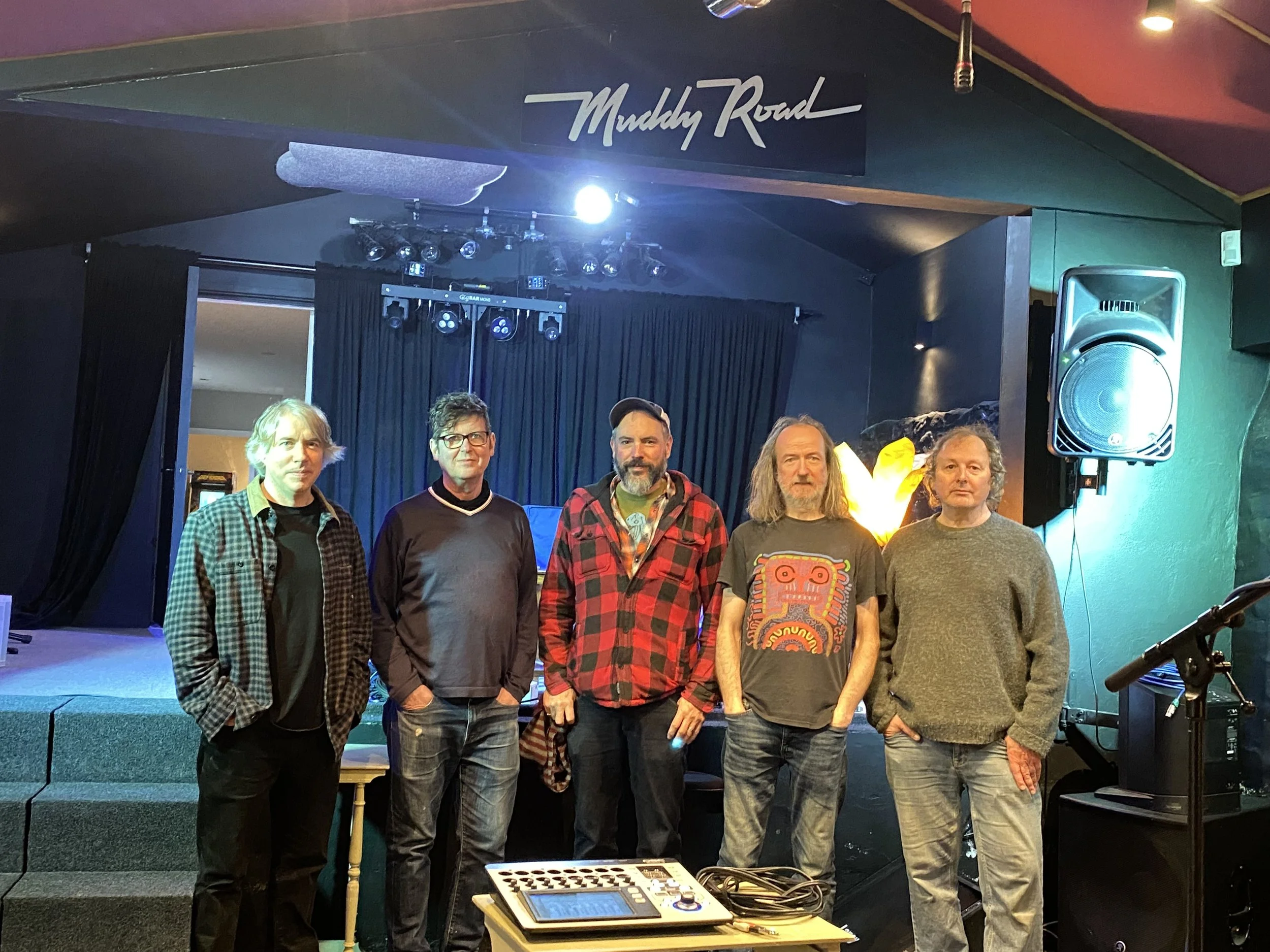 Five men stand on a stage in front of a black curtain with a sign that says 'Muddy Road.' The stage area includes lighting equipment and large speakers. There is a small mixing console in the foreground.