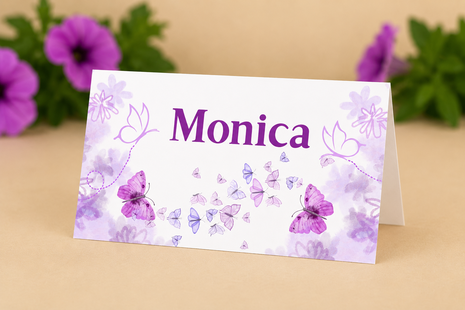 A place card with the name 'Monica' printed in purple, decorated with purple and pink butterflies and floral patterns, and placed on a beige table with purple flowers in the background.