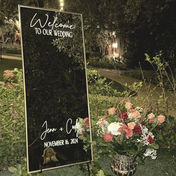 Wedding welcome sign with floral decoration at night, reading 'Welcome to our wedding, Jenn + Cody, November 16, 2024'.