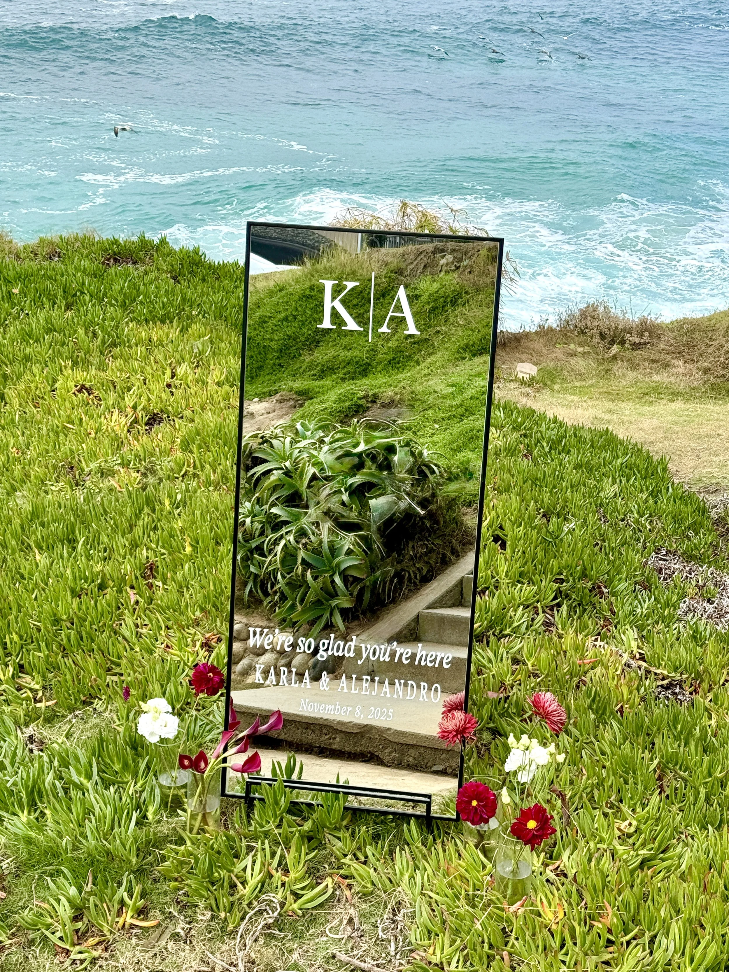 A wedding sign with the initials K and A, decorated with red and white flowers, placed on a grassy area near the ocean.