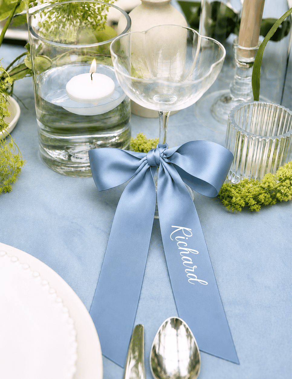 Personalized dusty blue satin ribbon place card on a champagne glass for an elegant wedding reception table setting.