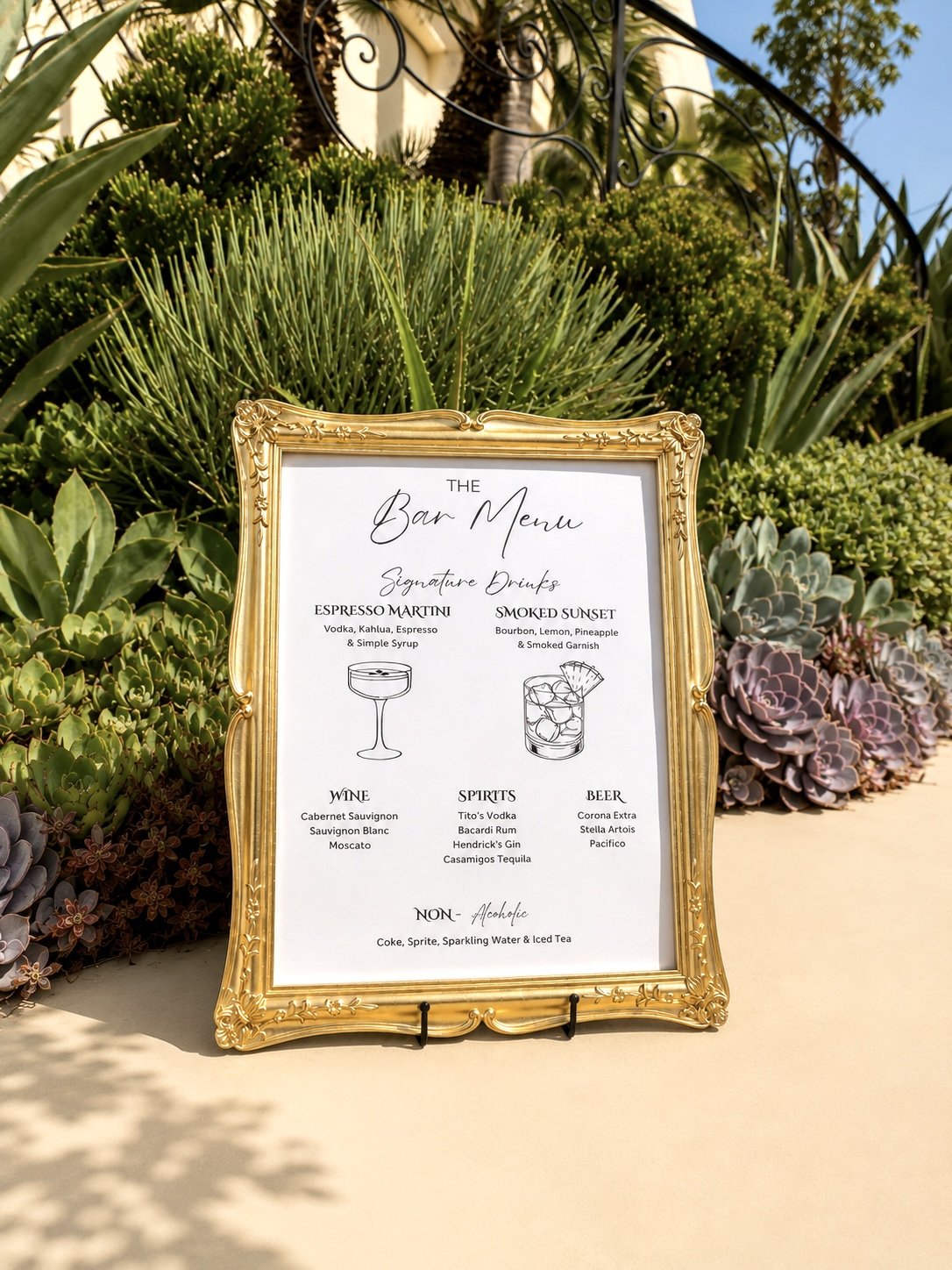 Custom wedding bar menu sign with modern calligraphy in a vintage gold frame, personalized with signature cocktails and displayed in a luxury garden wedding setting.