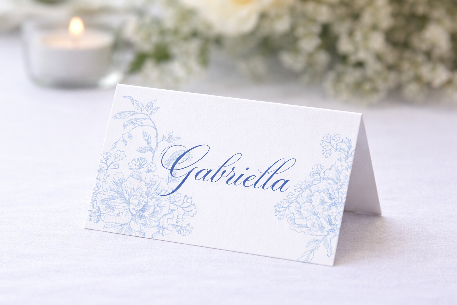 White folded place card featuring delicate blue floral illustrations and the name “Gabriella” in elegant cursive, displayed on a wedding reception table for assigned seating.
