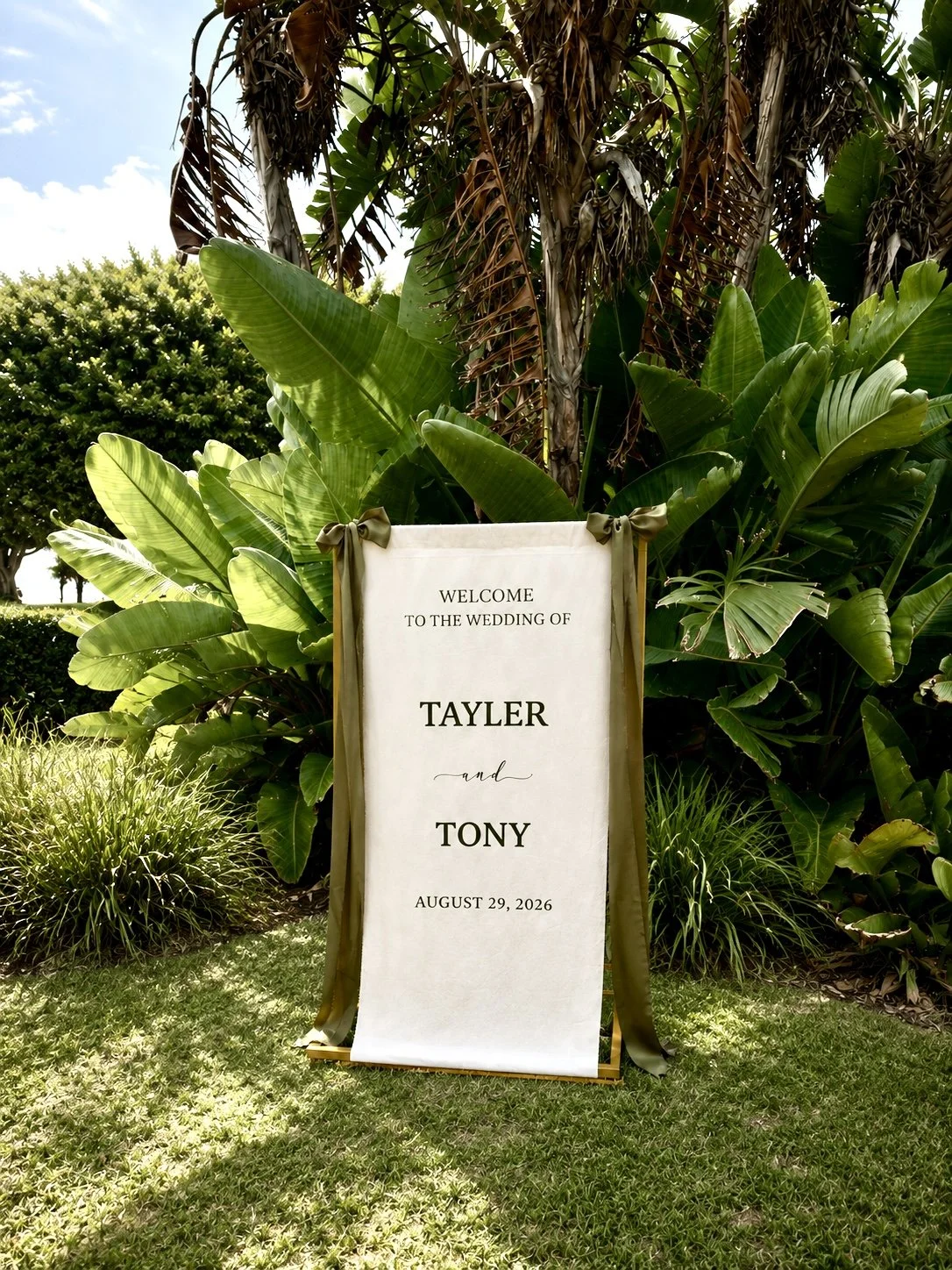 Welcome to the wedding of fabric sign with olive green font and satin ribbon displayed on a gold stand