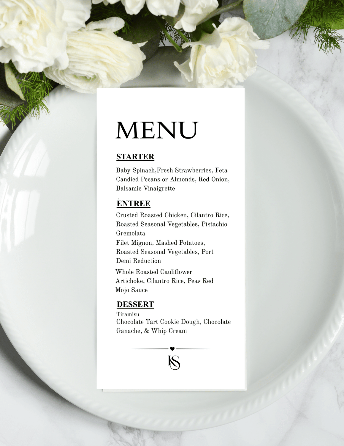 A white menu card with black text placed on a white plate, decorated with white flowers and green leaves.