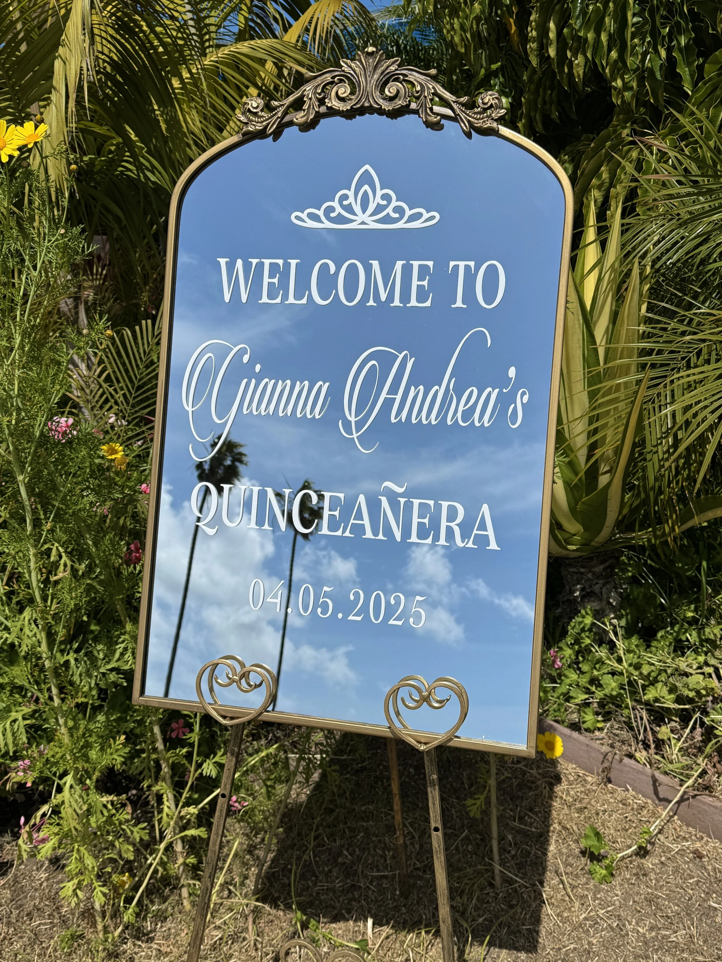 Ornate gold mirror welcome sign featuring a crown and personalized white lettering that reads, “Welcome to Gjianna Andrea’s Quinceañera 04.05.2025.