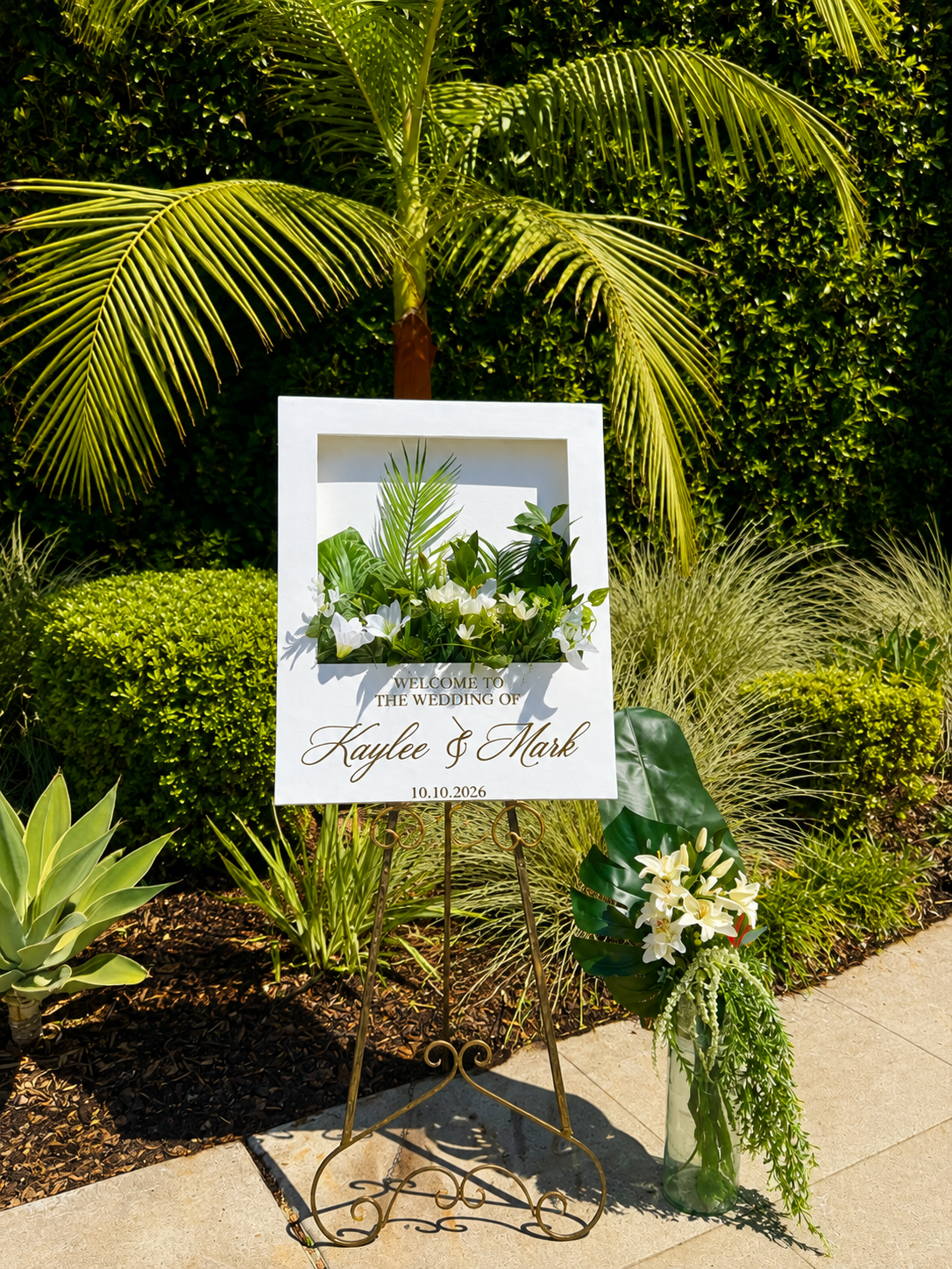 A wedding flower box welcome sign with white flowers and tropical greenery, on a metal easel