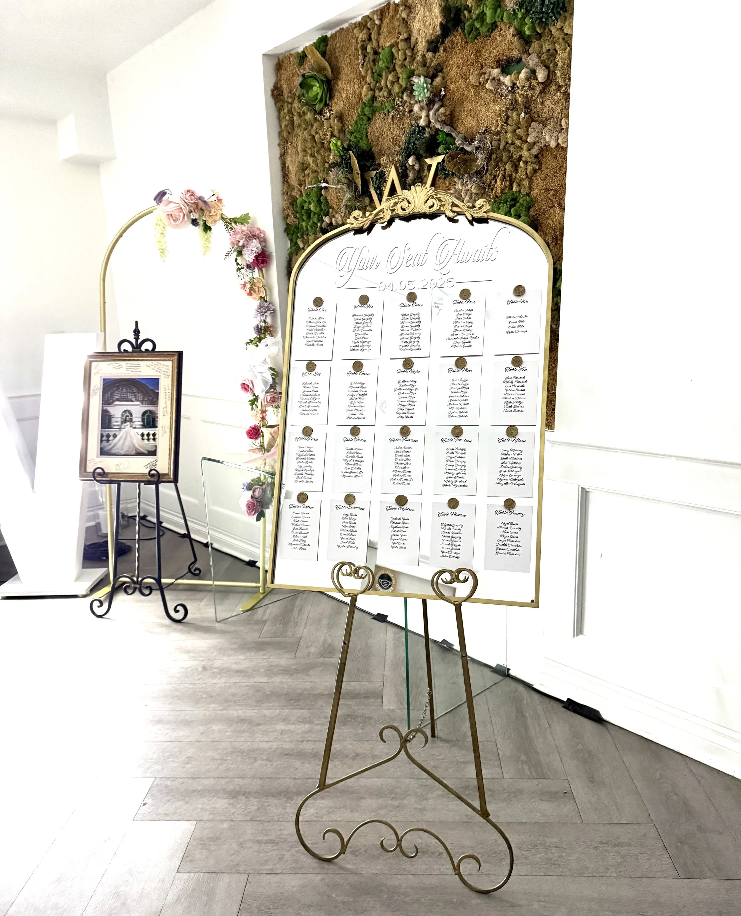 Quinceañera seating chart displayed on an ornate gold mirror, featuring white table-assignment cards with guest names and elegant gold wax seals.