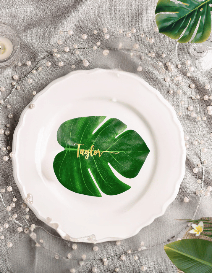 Large green monstera leaf personalized with the name “Taylor” in gold lettering, styled as a tropical wedding reception place card.