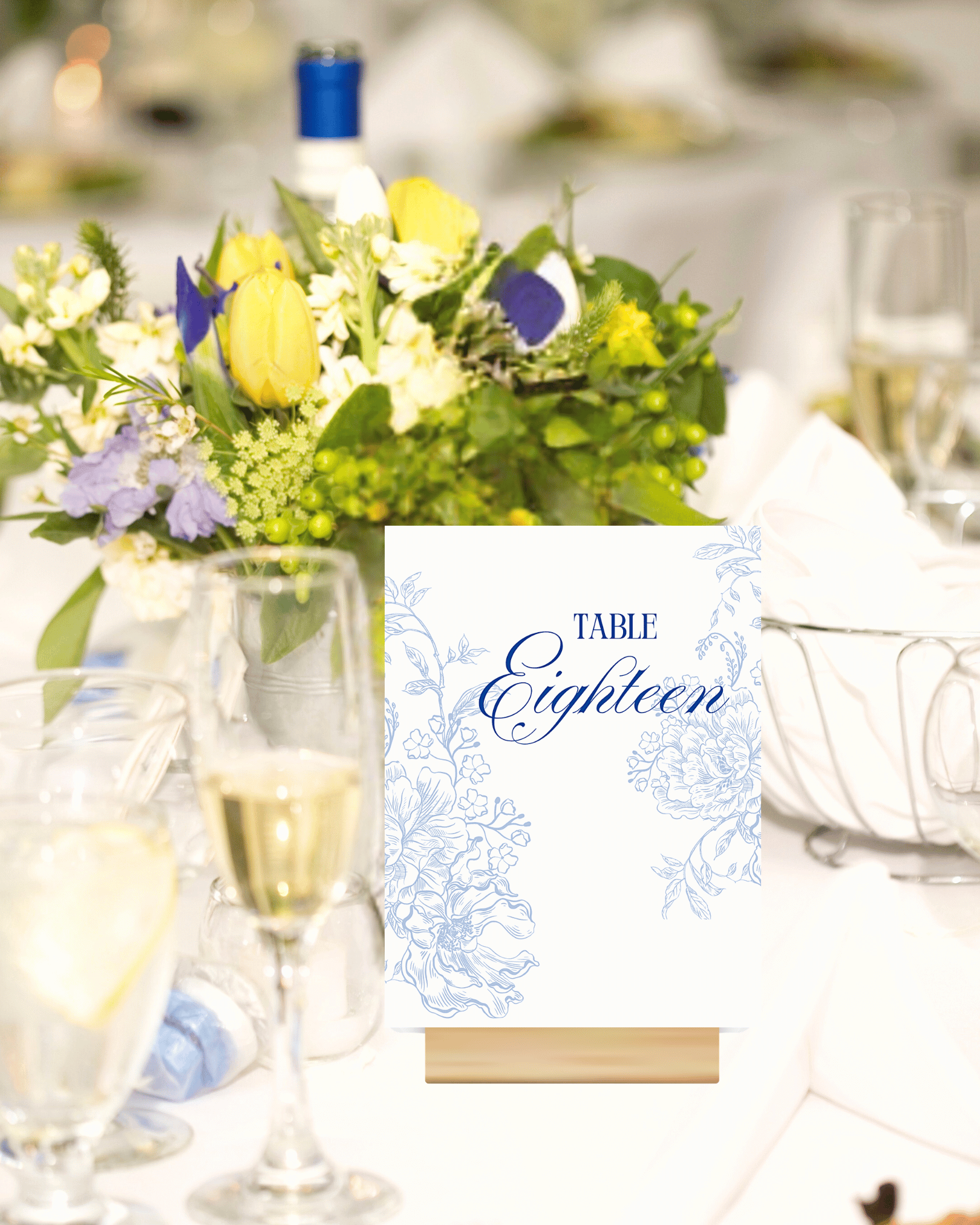 A floral centerpiece with yellow, purple, and white flowers on a table at a wedding reception, along with champagne glasses and a seating chart with the word 'Eighteen' visible.