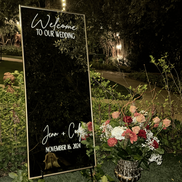 A tall, vertically oriented black mirror sign with white cursive and block text welcoming guests to a wedding on November 16, 2024, and displaying the names Jenn and Cody. The sign is positioned outdoors next to a floral arrangement with pink, white, and red flowers, and greenery, at night time.