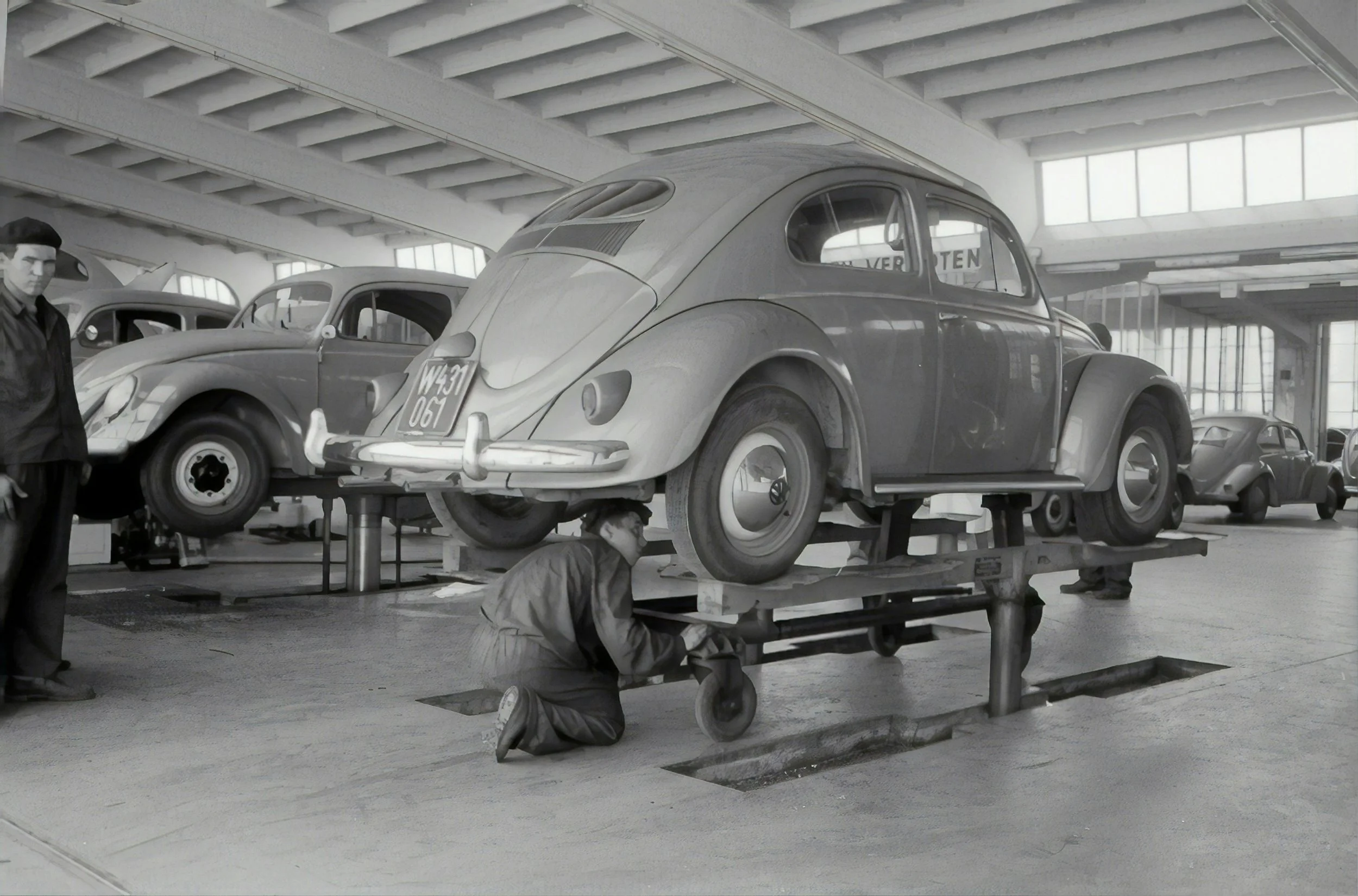 Black and white photo of vintage cars being assembled in an auto factory with workers inspecting and working on the vehicles.