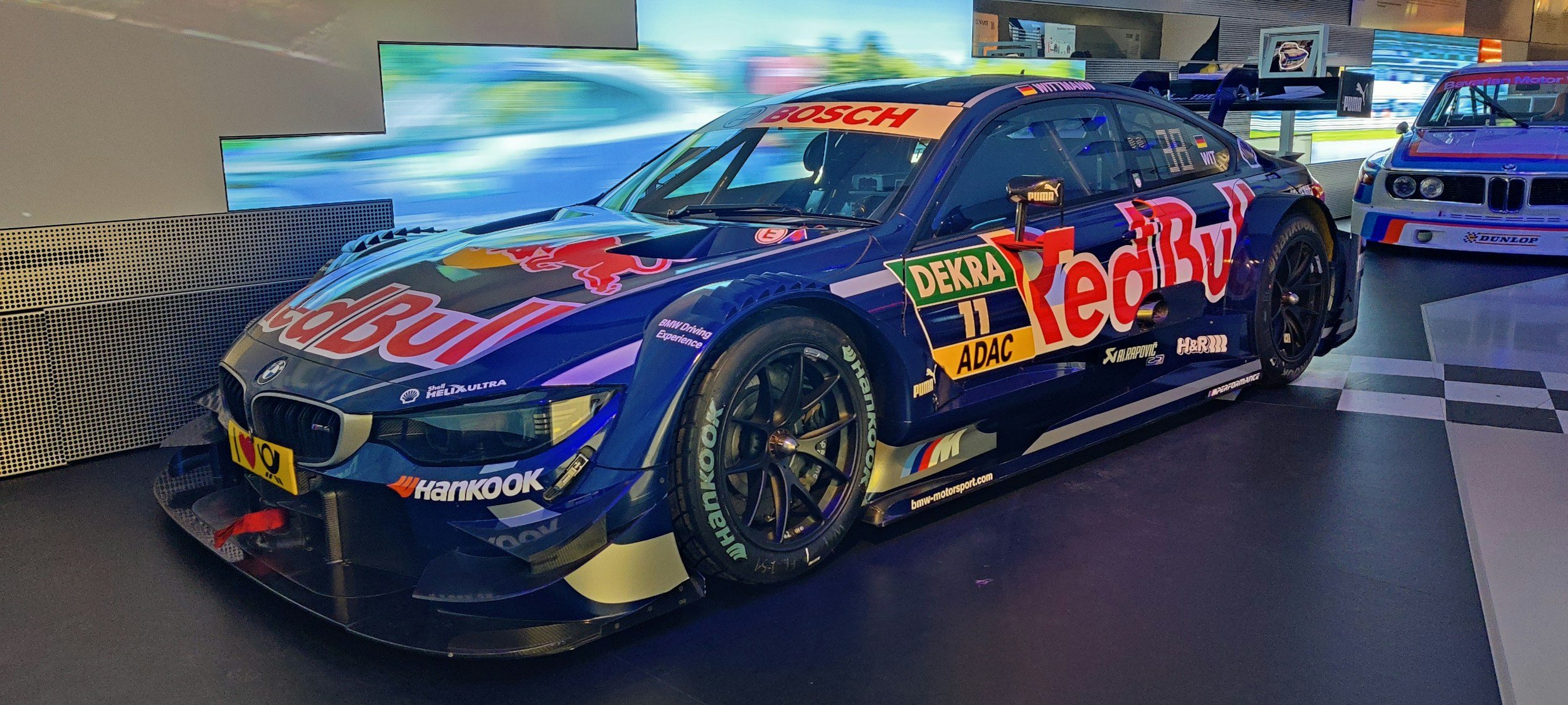 Blue BMW race car with Red Bull and sponsor logos on display indoors, next to another race car.