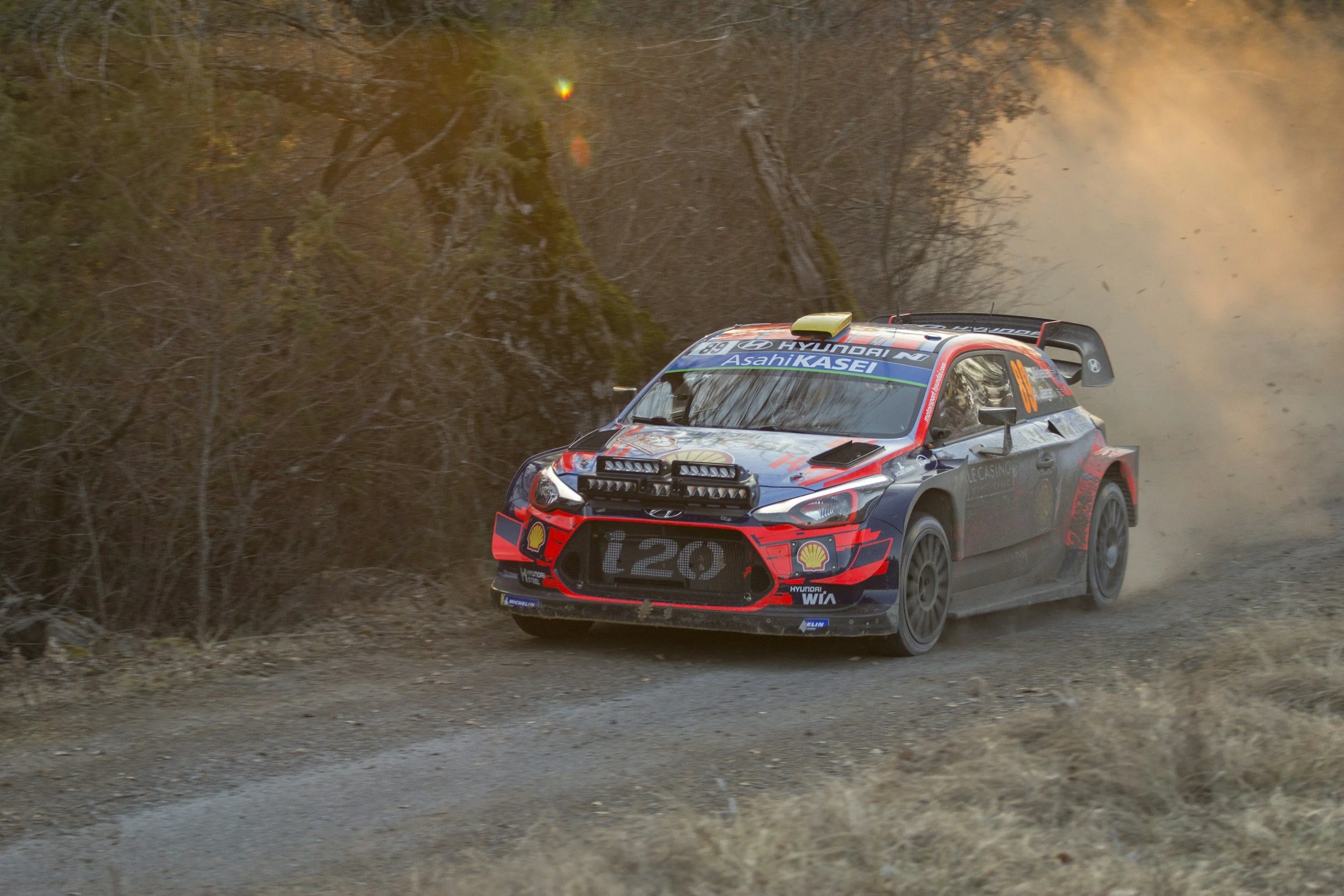 A rally car racing on a dirt road through a wooded area, kicking up dust behind it.