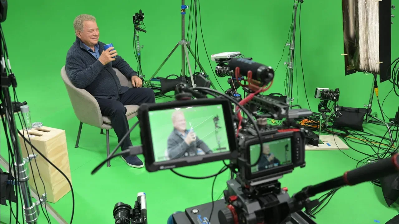 An older man sitting in a studio with a green screen background, holding a blue cup and smiling, during a filming or photoshoot session with professional camera and lighting equipment around.