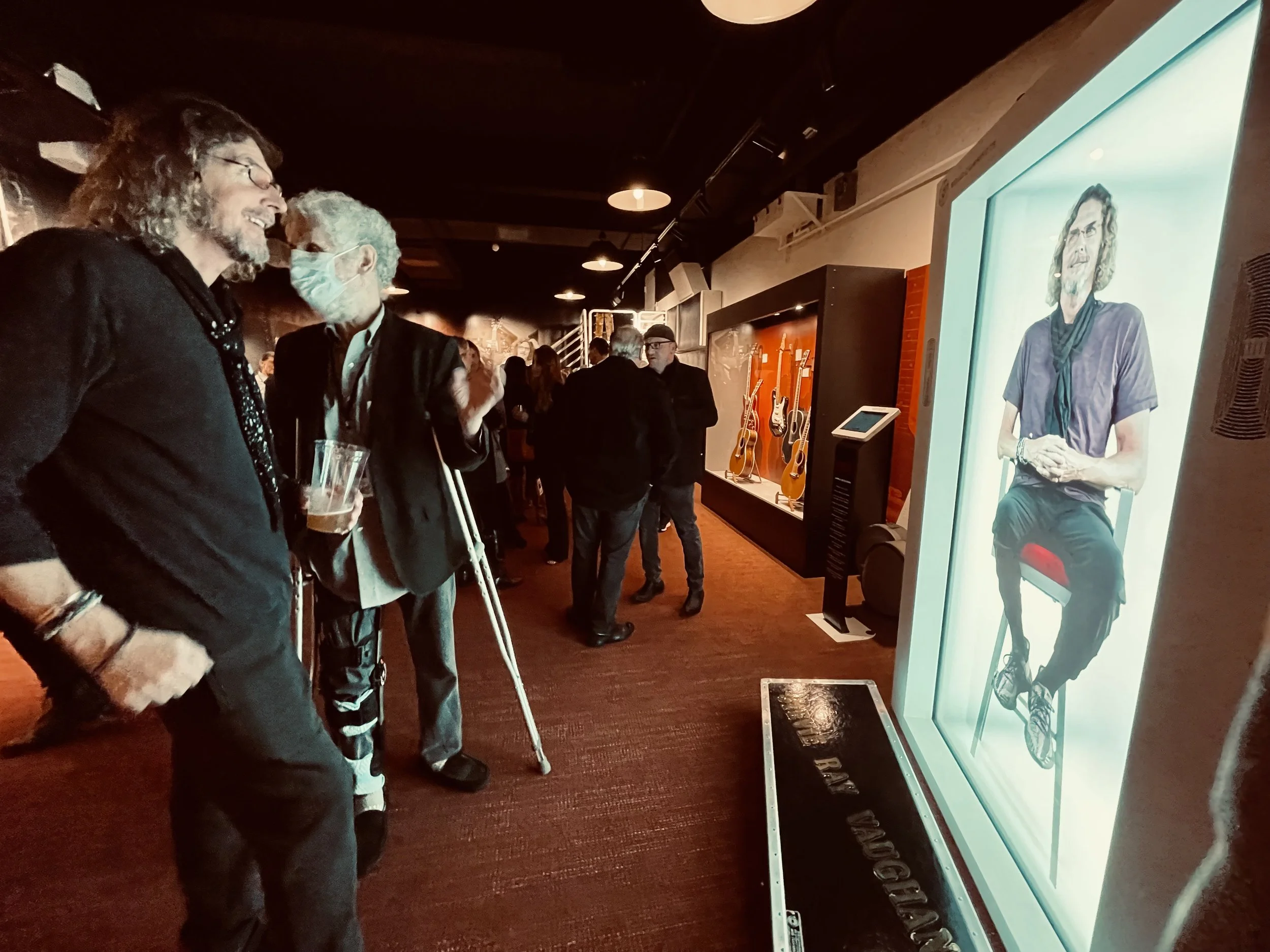 People at an art exhibit looking at a digital artwork of a man sitting in a chair with long curly hair and glasses, displaying on a large screen.