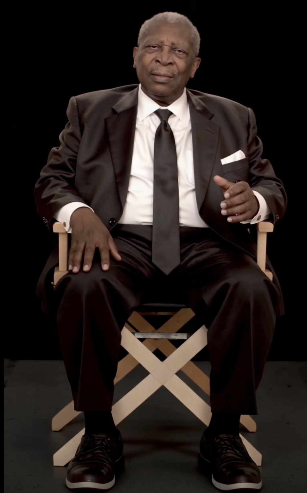 An elderly man in a black suit, white shirt, and black tie sitting on a director's chair against a black background.