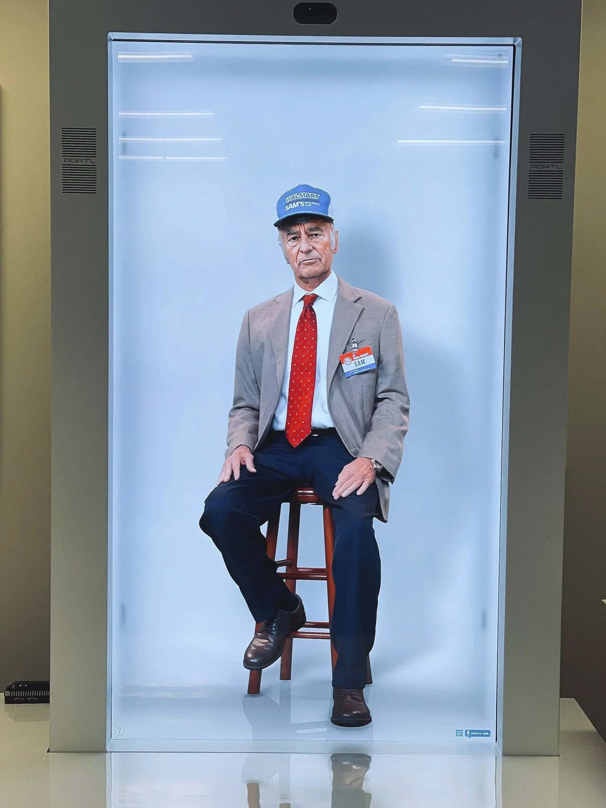 A digital advertisement display shows a man sitting on a stool, wearing a beige blazer, white shirt, red tie, and a blue cap with Walmart and Sam's Club logos. The man appears serious and is looking directly at the camera.