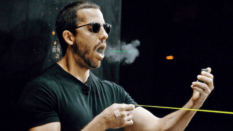 A man with sunglasses exhales smoke while holding a yellow laser string in his hands.