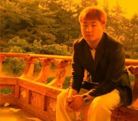 Young man sitting outdoors on a wooden railing during sunset, wearing a black jacket and white pants.