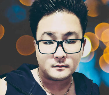 A man with black glasses and dark hair, wearing a dark jacket, against a bokeh background of colorful lights.