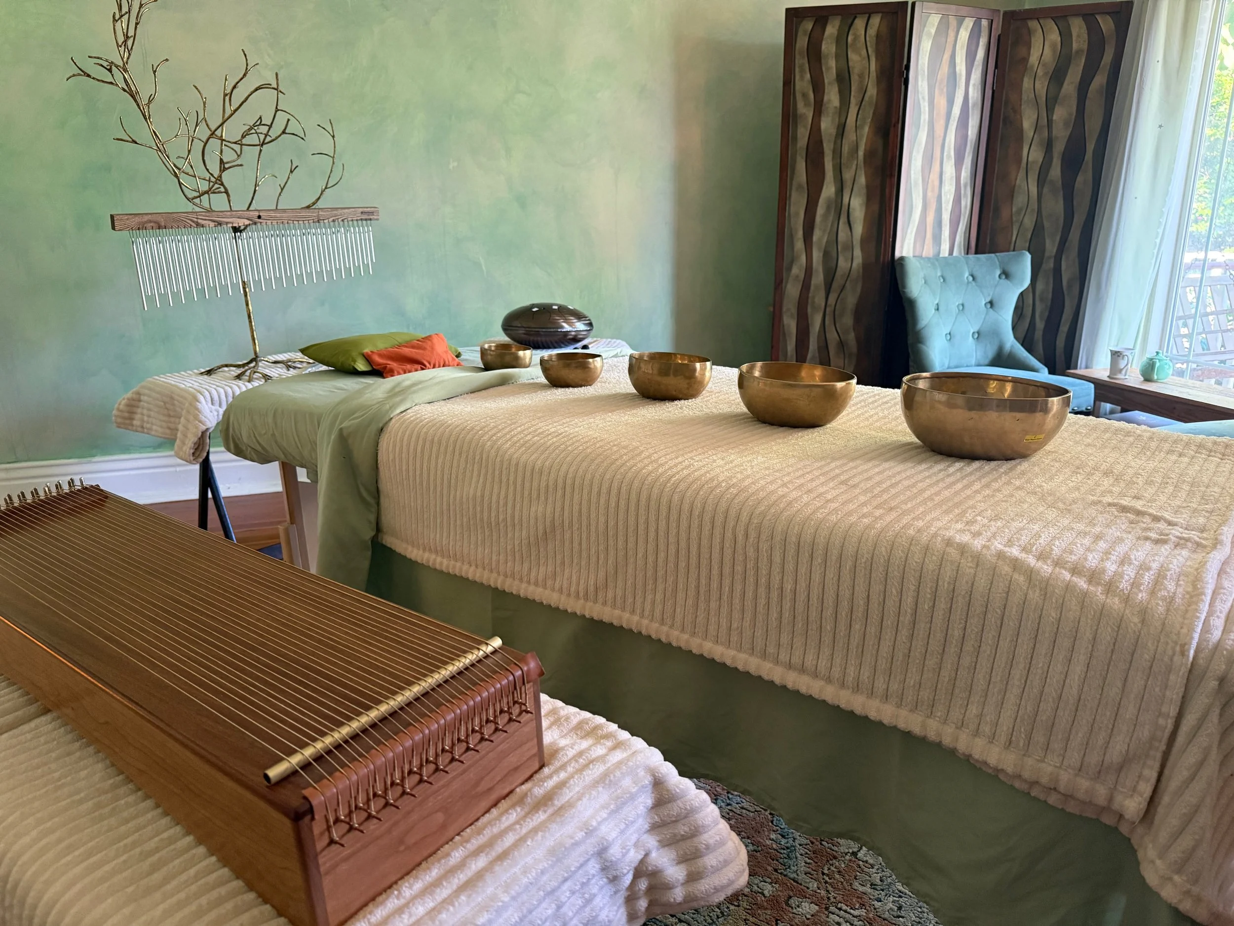 A room arranged for a crystal healing or massage therapy session with a massage table at the center, draped in a beige blanket and towels. Surrounding tables hold bowls and instruments, with herbal plants and decorative items on shelves. The walls are painted in light green, and natural light illuminates the space from a nearby window.