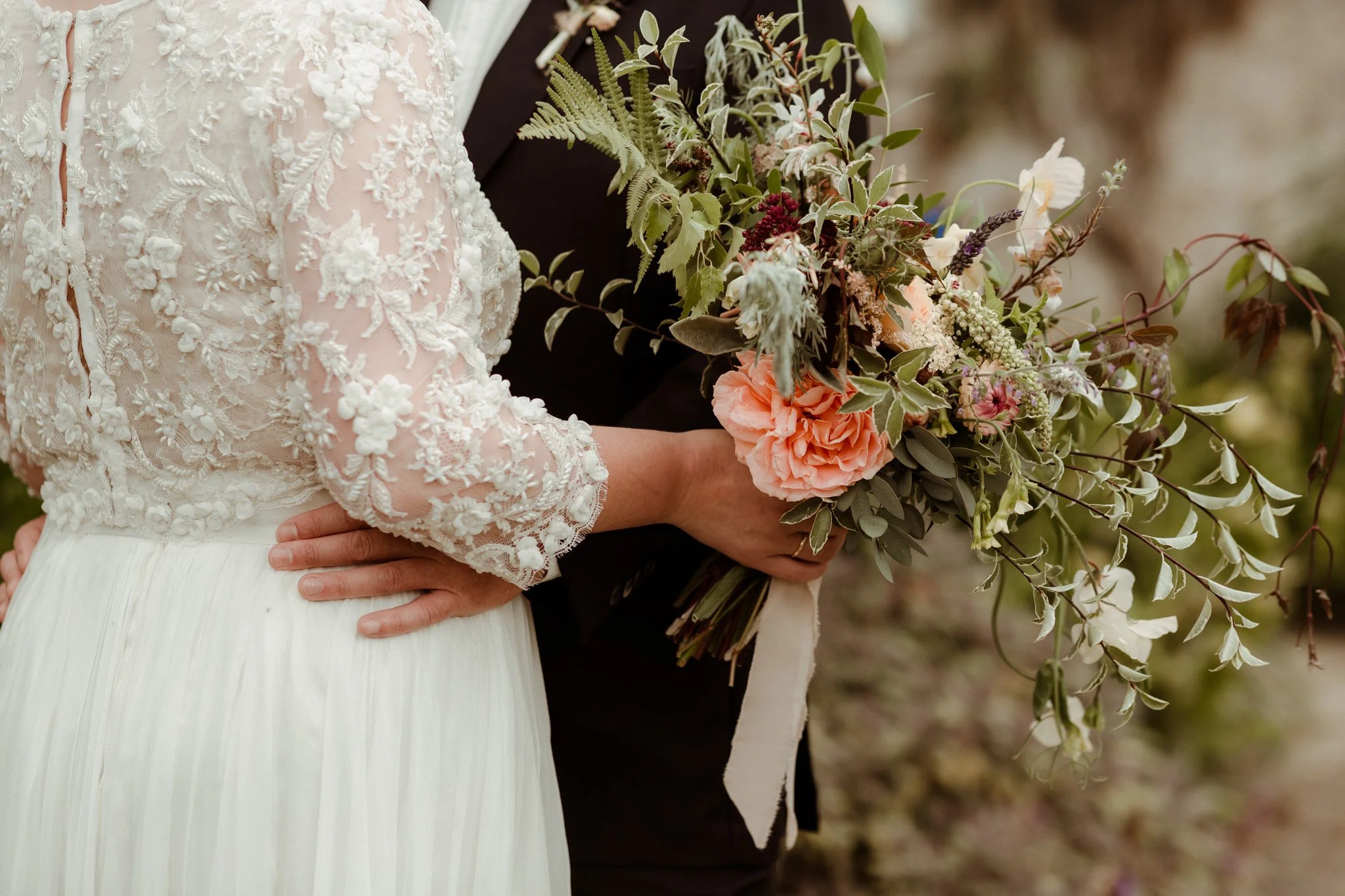 Classic bride in white lace dress holding bespoke organic Devon-grown natural flower bouquet whilst embracing groom in dark suit