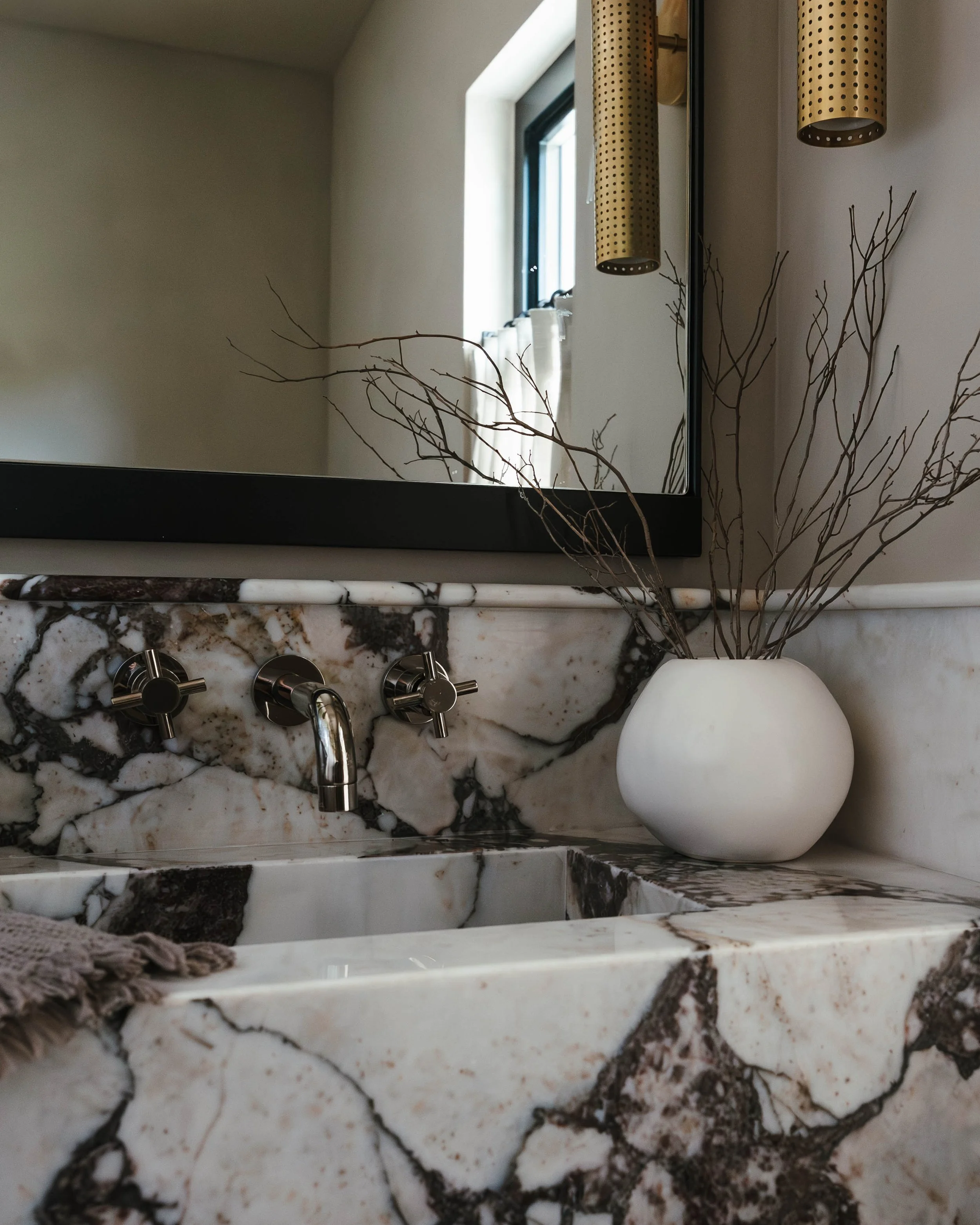 A bathroom sink with a marble countertop, a white vase with bare branches, and a mirror reflected window with a beige curtain.