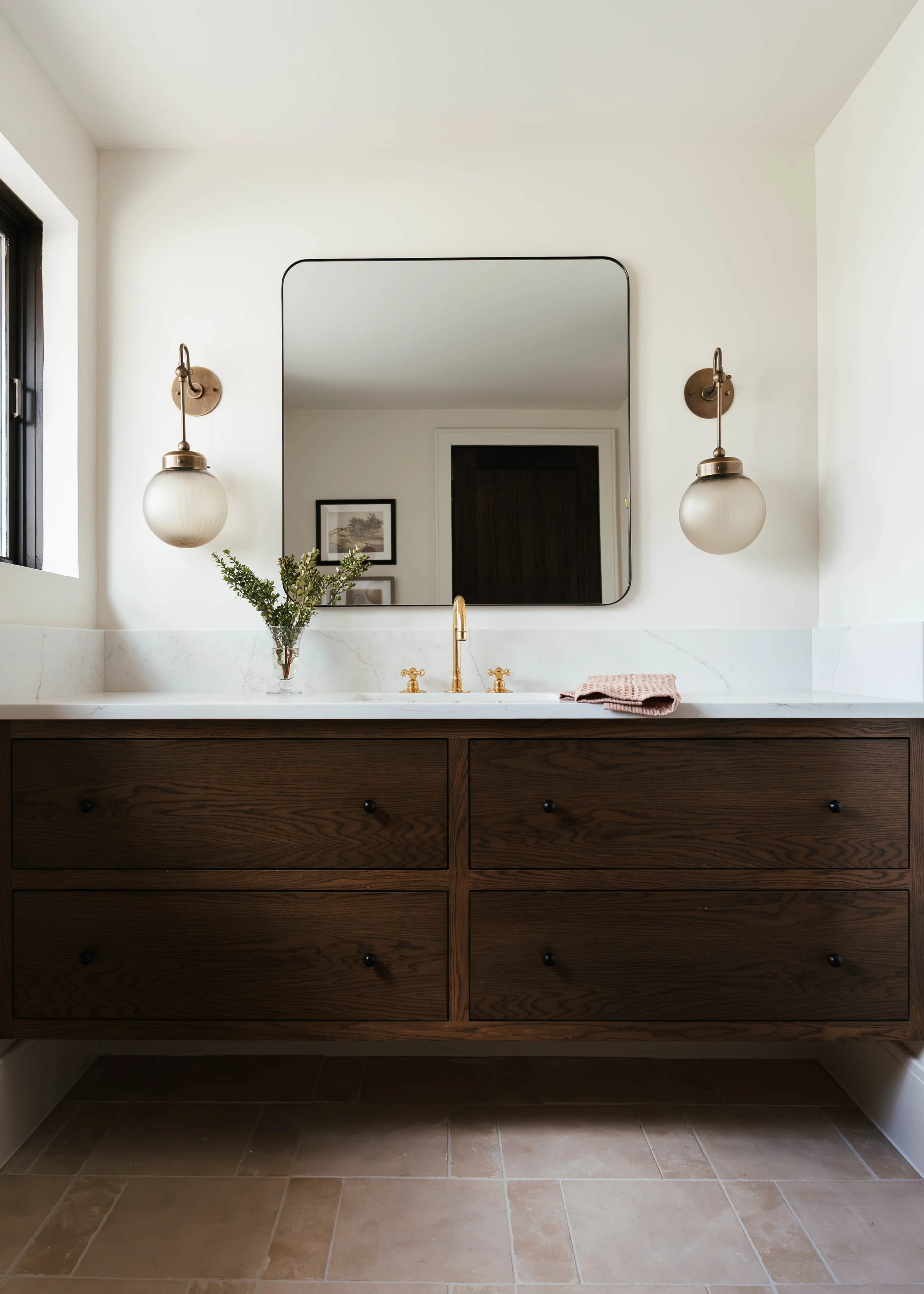 Bathroom vanity with dark wood drawers, a gold faucet, and a marble countertop, decorated with a small plant, a pink towel, a mirror, and wall-mounted lights on either side.
