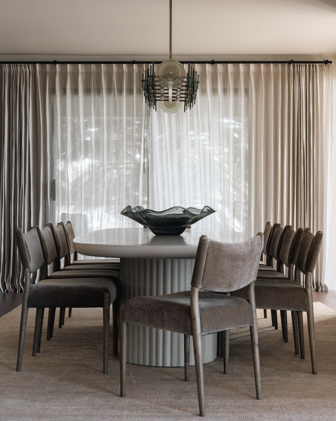 Dining room with a large white round table, eight upholstered chairs, a black decorative bowl on the table, beige curtains, and a modern chandelier hanging from the ceiling.