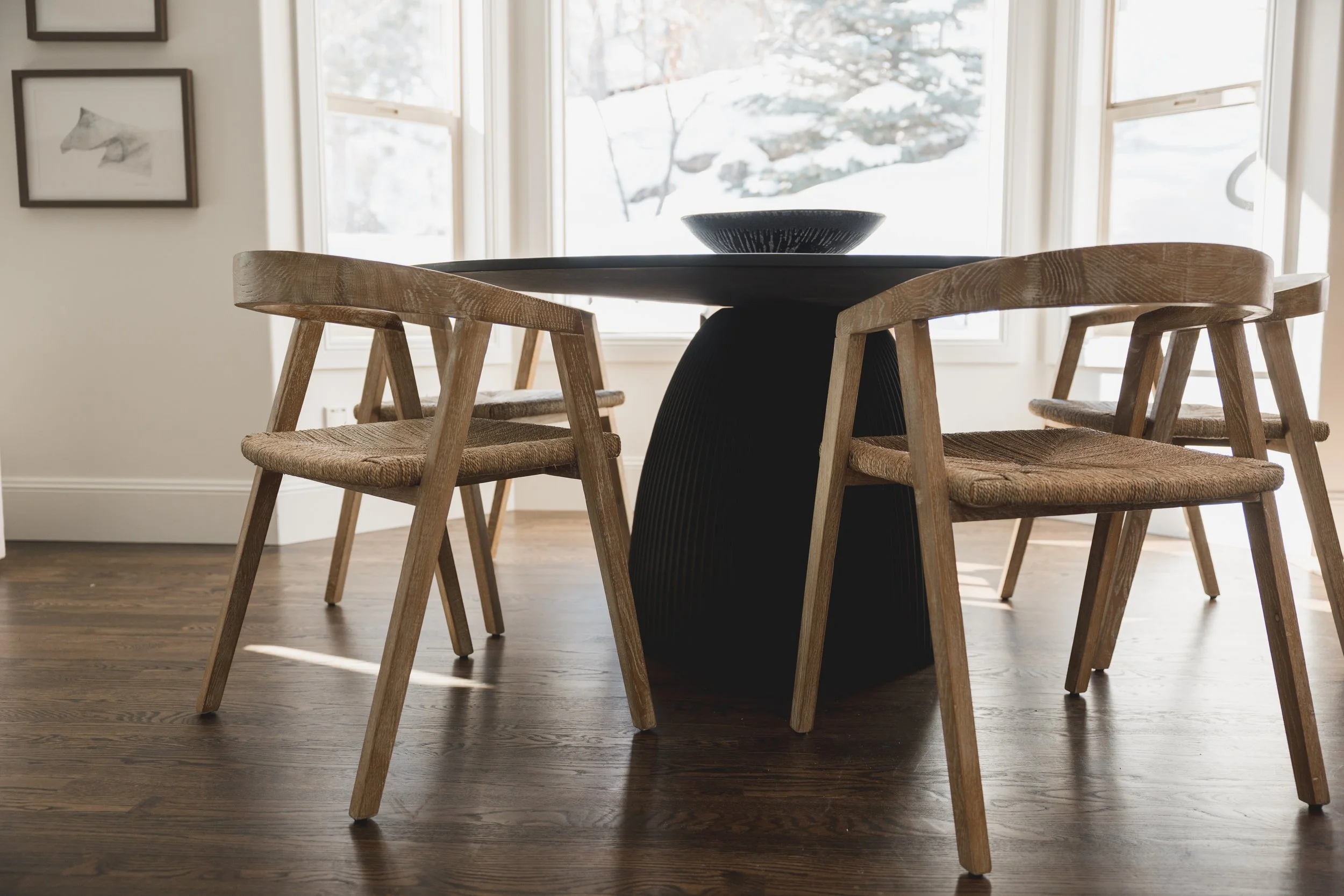 Wooden dining table with four matching wooden chairs, in a bright room with large windows and framed artwork on the walls.