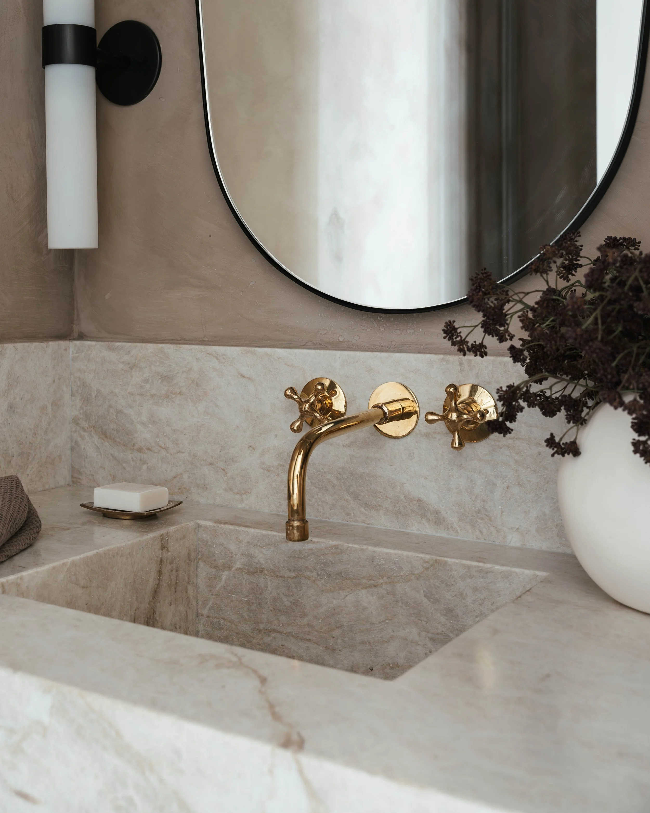 A bathroom sink with a gold faucet, soap on a tray, a gray towel, a round mirror, a modern wall sconce, and a white vase with dark purple flowers.