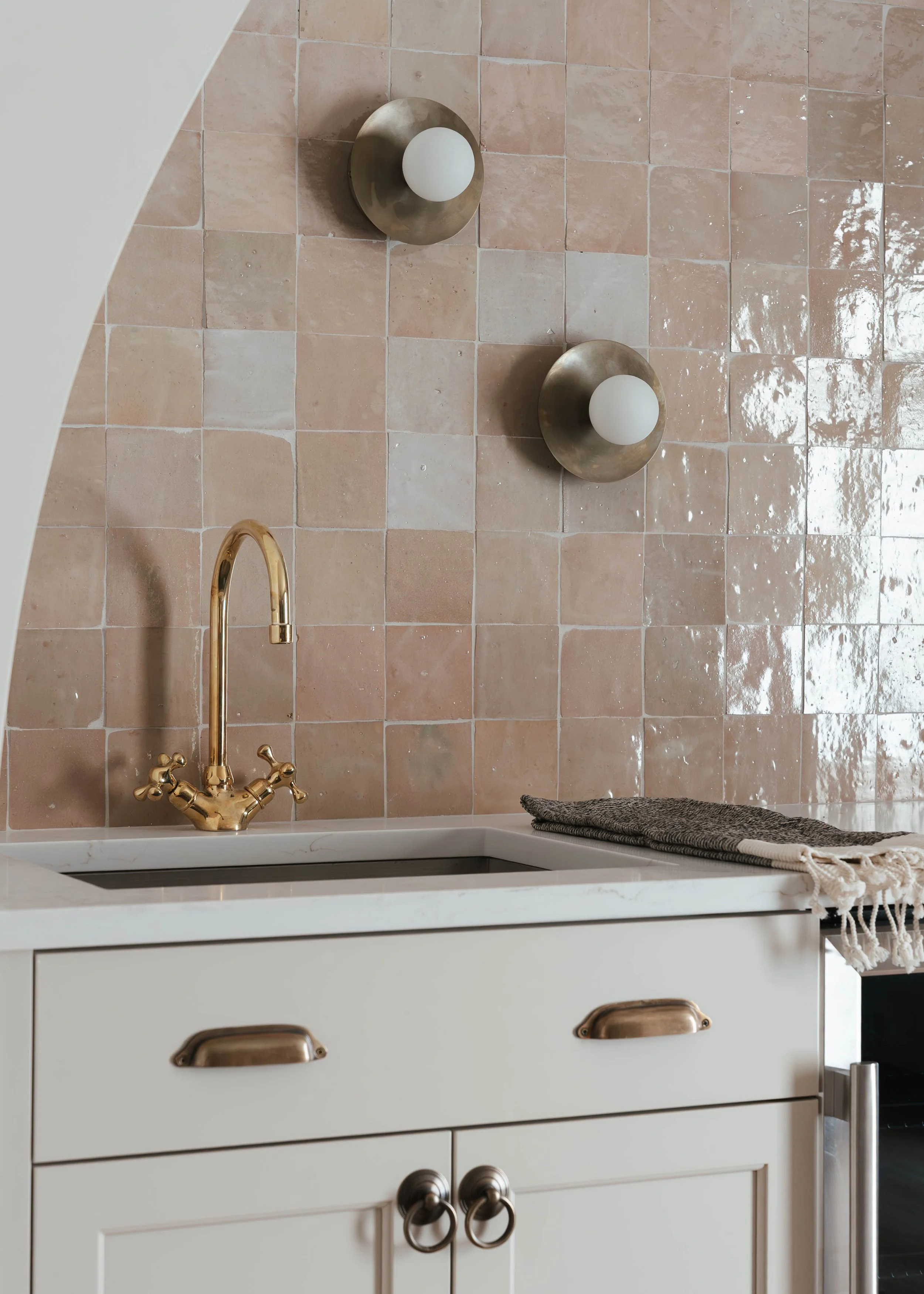 A kitchen with a beige tile backsplash, a gold faucet, and a white cabinet with brass handles. There are two round wall sconces with frosted globes on the wall, and a gray cloth on the countertop.