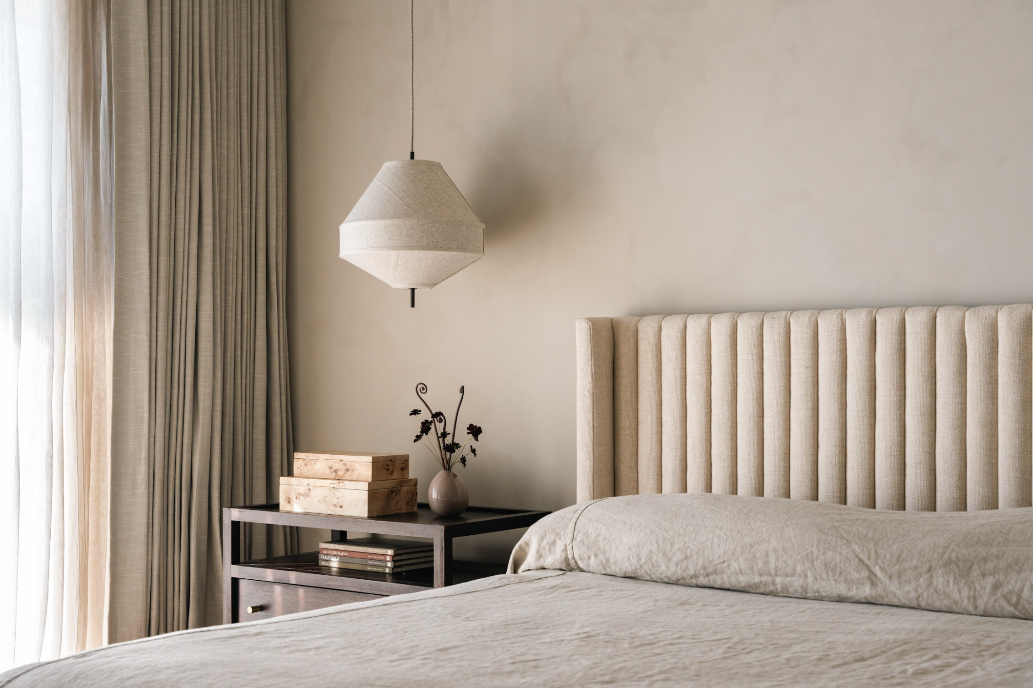A bedroom with beige walls, a cream-colored headboard, a bed with light-colored bedding, and a nightstand with decorative boxes, a vase with flowers, and an overhead hanging light.